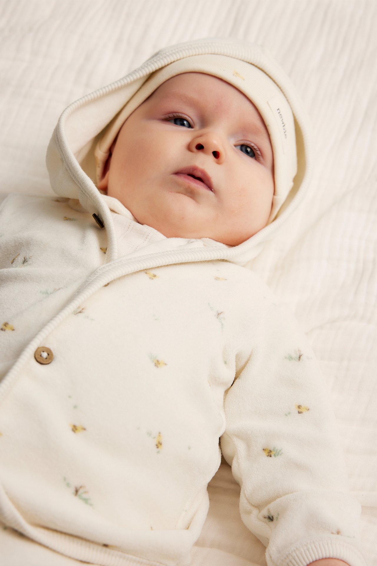 Frontvisning av en baby i en offwhite mønstret velourhettegenser med treknapper og en matchende lue.