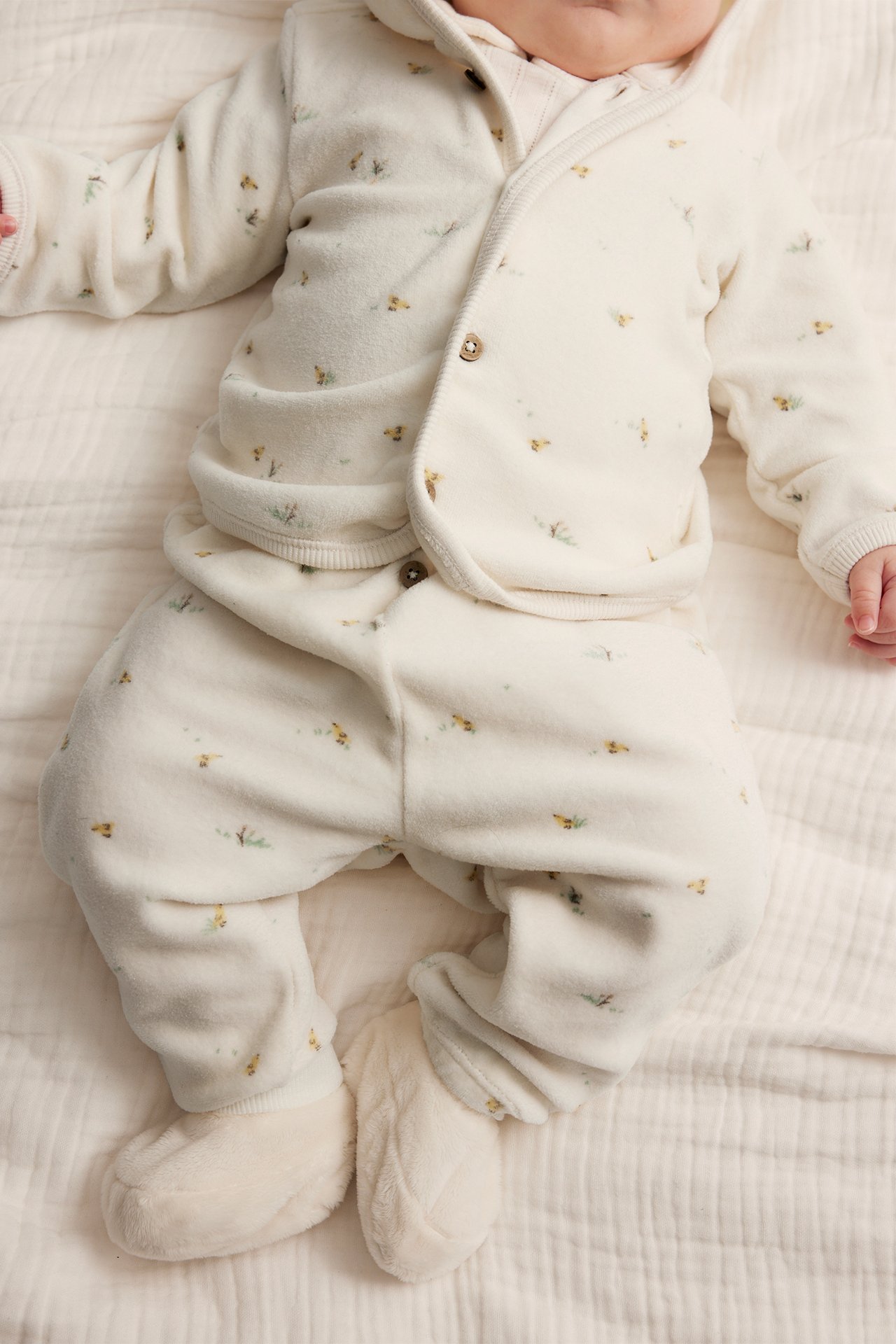 Ovenfra-bilde av baby i offwhite mønstret velourhettejakke med knapper, matchende bukser og myke tøfler.