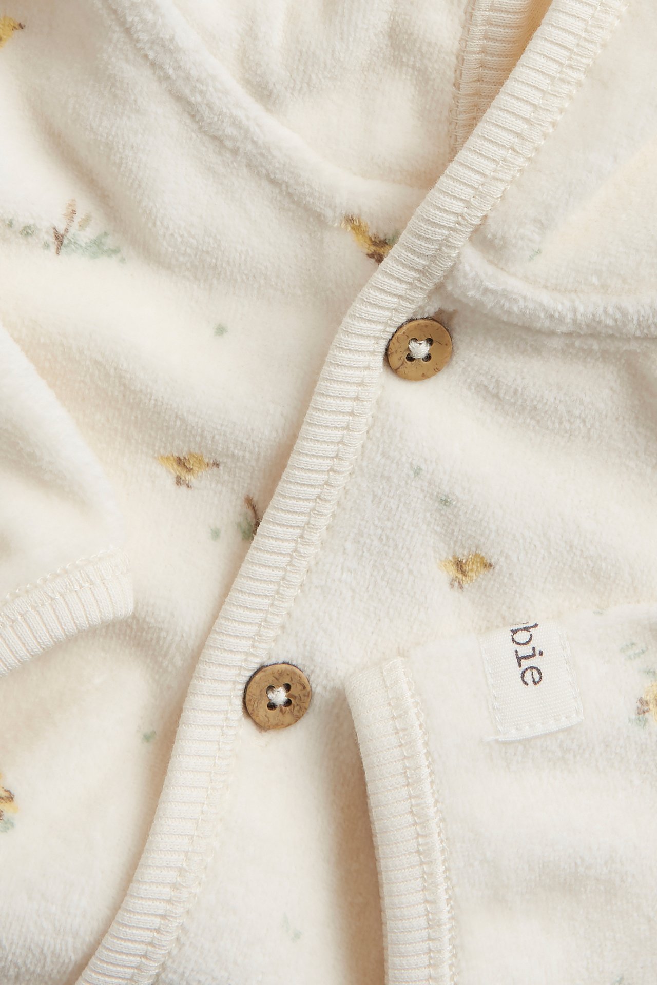 Nærbilde av en offwhite mønstret velour hettegenser for baby med treknapper foran.