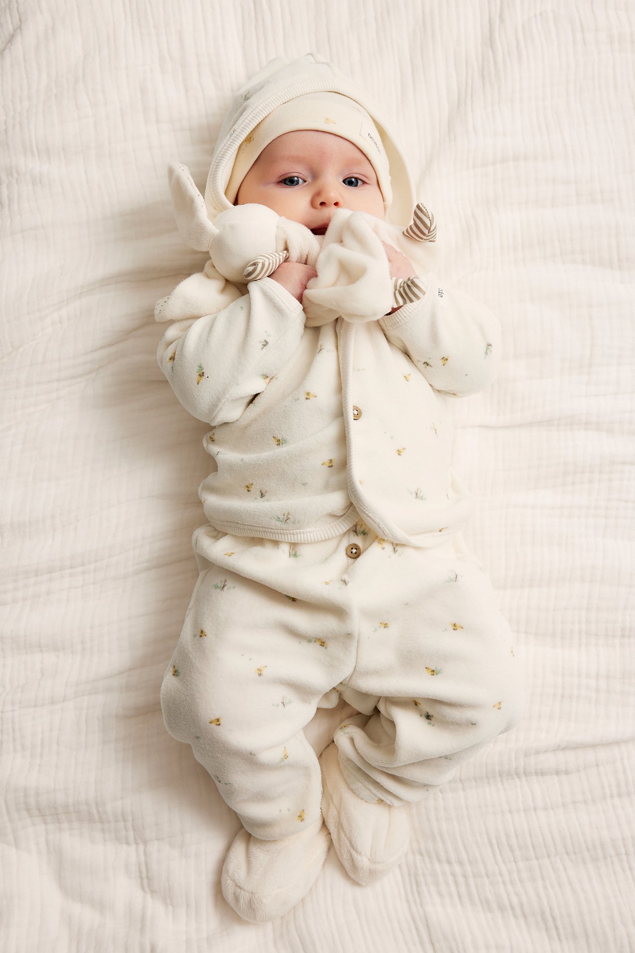 Ovenfra: Baby i offwhite mønstret velour hettegenser, matchende bukser, lue, tøfler, holder en leke.