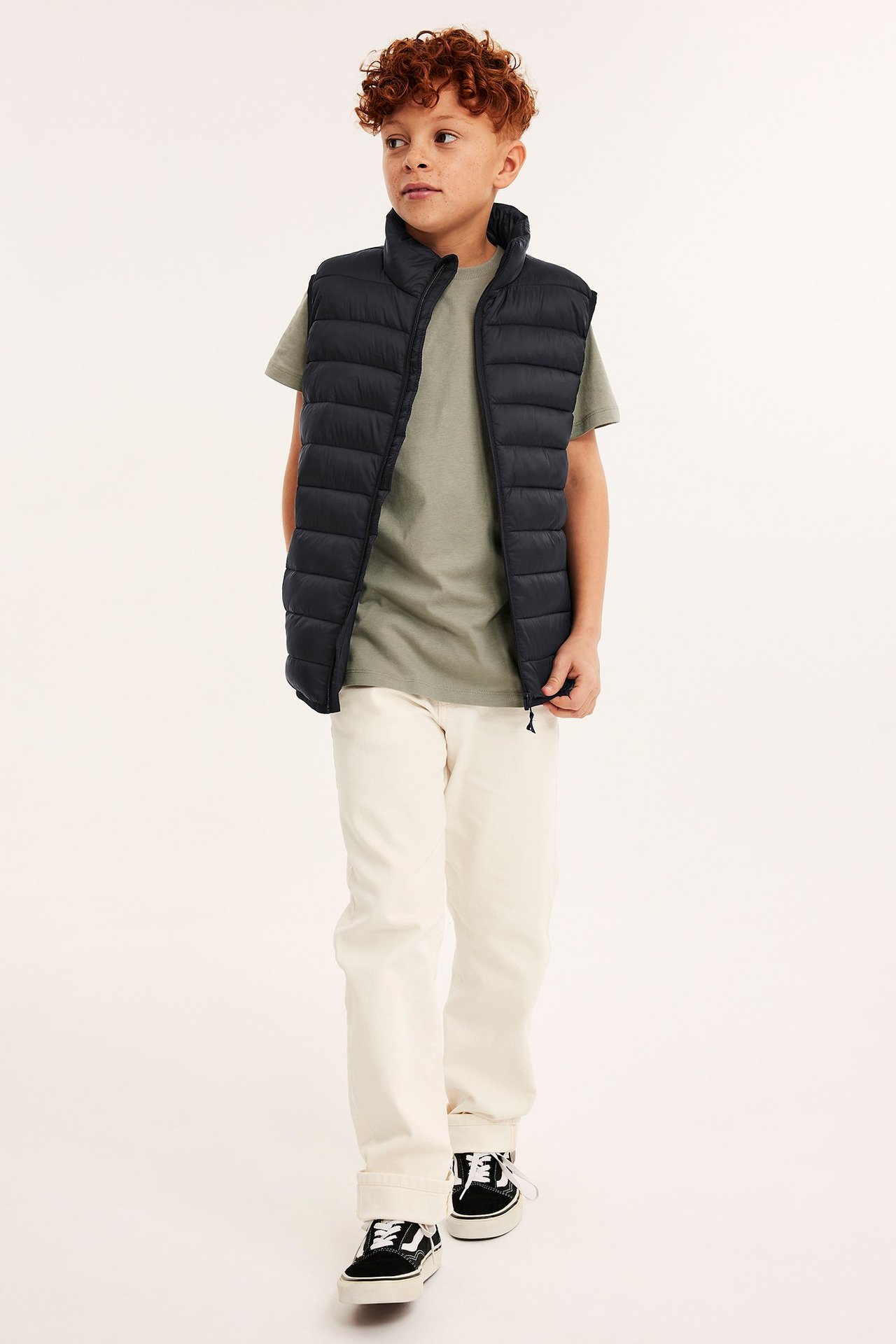 Frontvy av ett barn i svart vadderad lättviktsväst över en olivgrön t-shirt, gräddvita byxor och svarta sneakers.