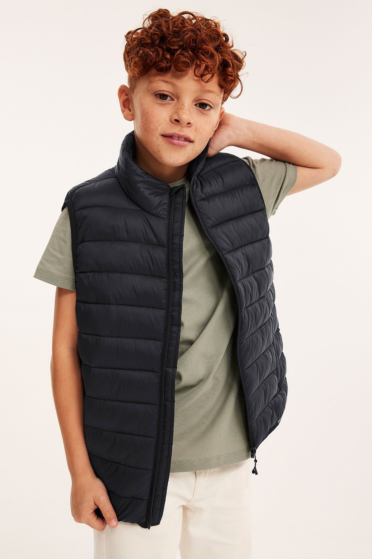 Widok z przodu dziecka w otwartej czarnej pikowanej lekkiej kamizelce na zielonej koszulce i jasnobeżowych spodniach.