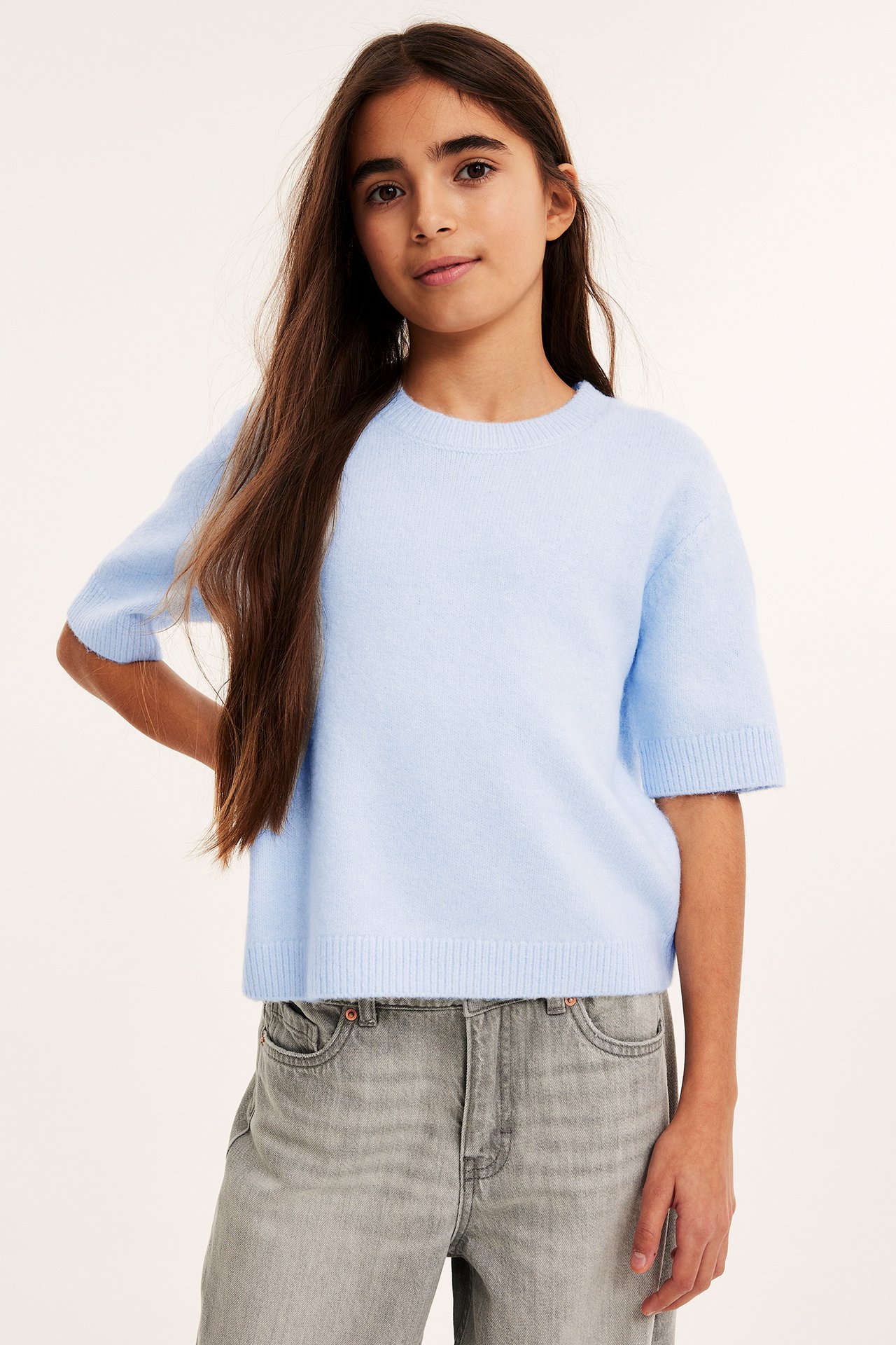 Forfra, et barn i en lyseblå kortermet finstrikket genser og grå jeans.