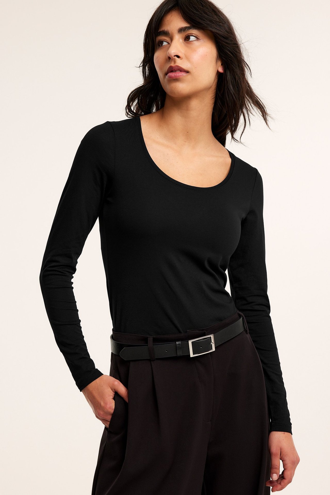 Forfra, kvinne i svart langermet topp med scoop-hals, mørke bukser og svart belte.