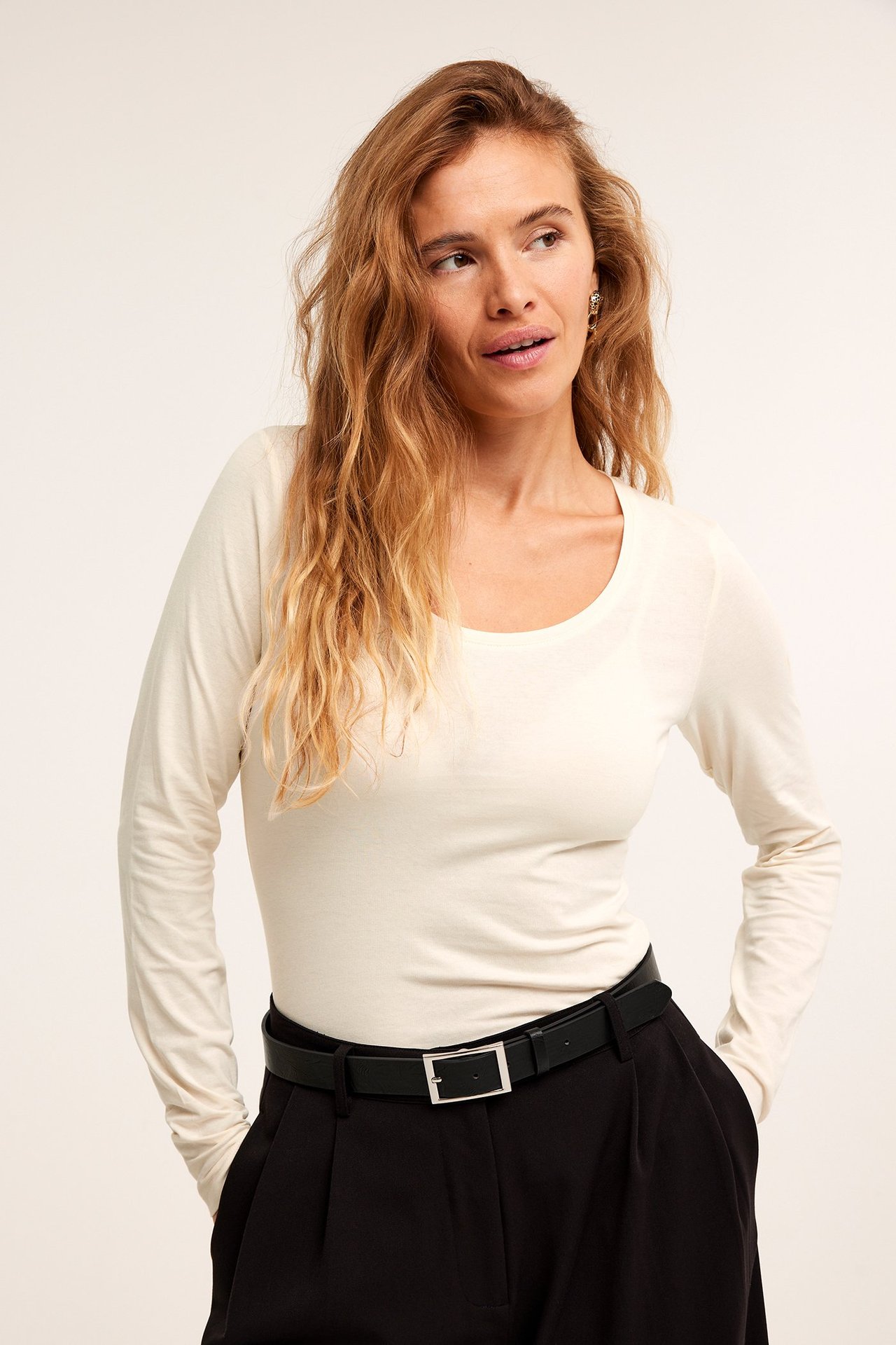 Frontbilde av en kvinne i offwhite langermet topp med scoop-hals, svarte bukser og et svart belte.