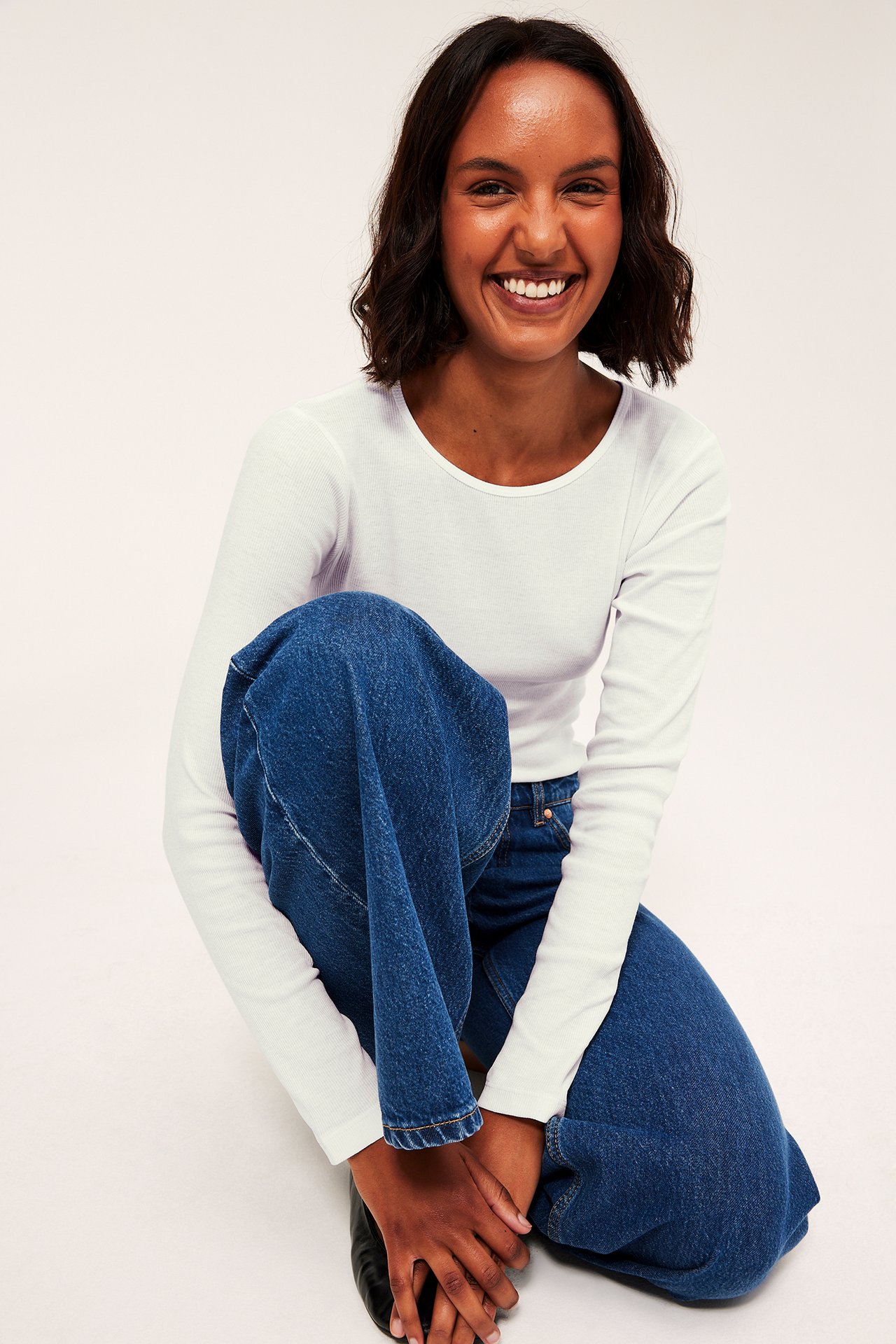 Frontvy av en kvinna i en vit långärmad ribbad topp och blå vida jeans.