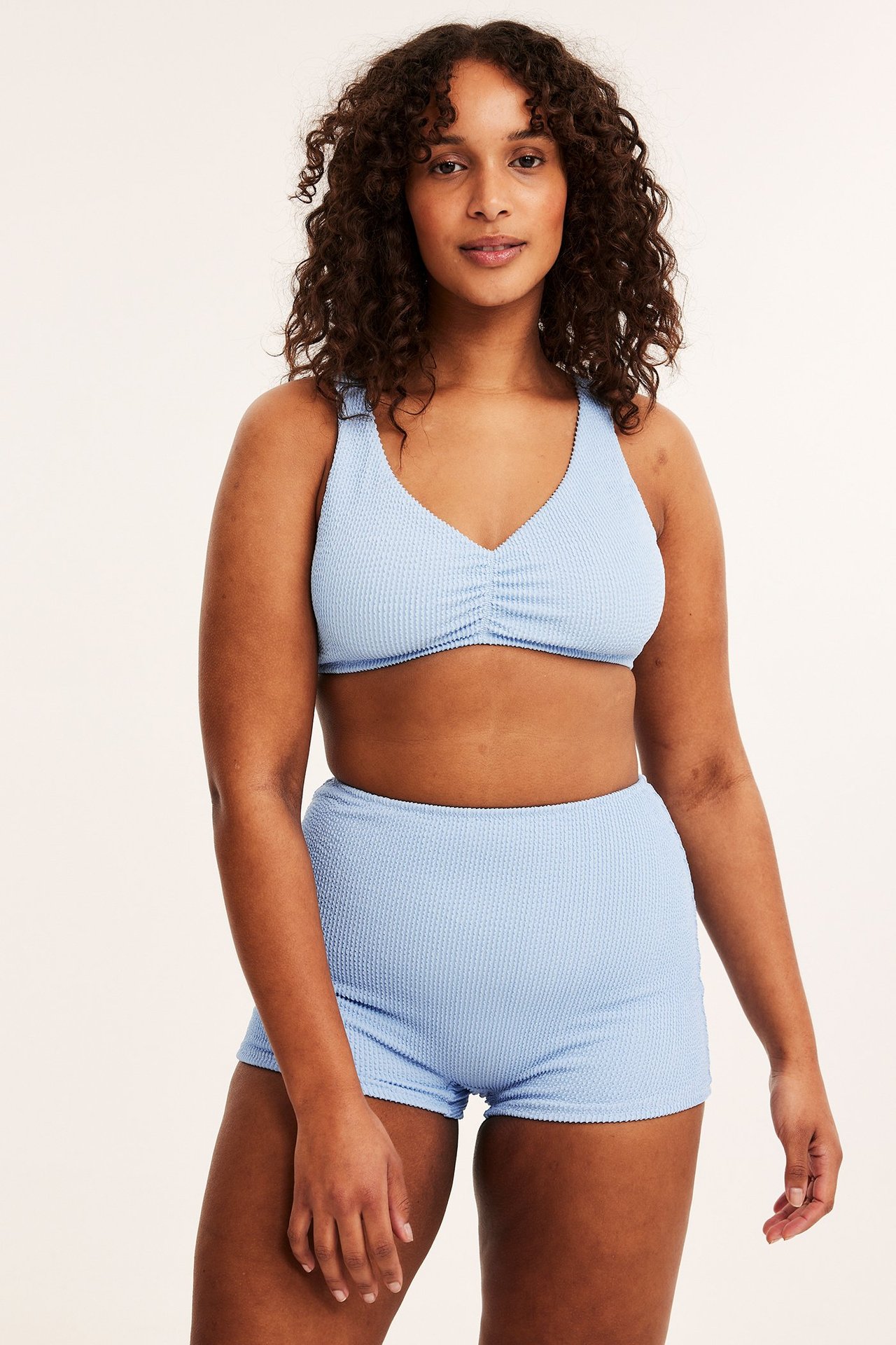 Frontbilde av kvinne i lyseblå, høytlivet, strukturert bikinishorts og matchende V-hals bikinitopp.