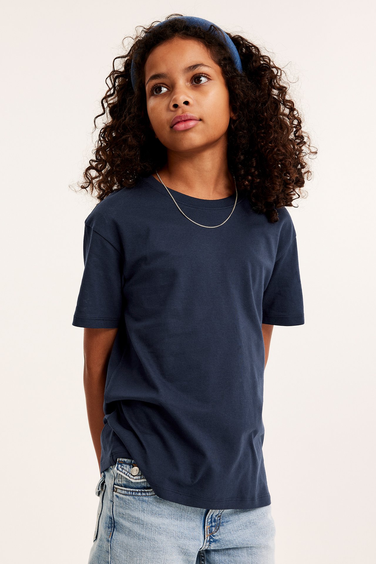 Framsida av ett barn i en mörkblå bomulls-t-shirt, ljusblå denimshorts, blått hårband och silverhalsband.