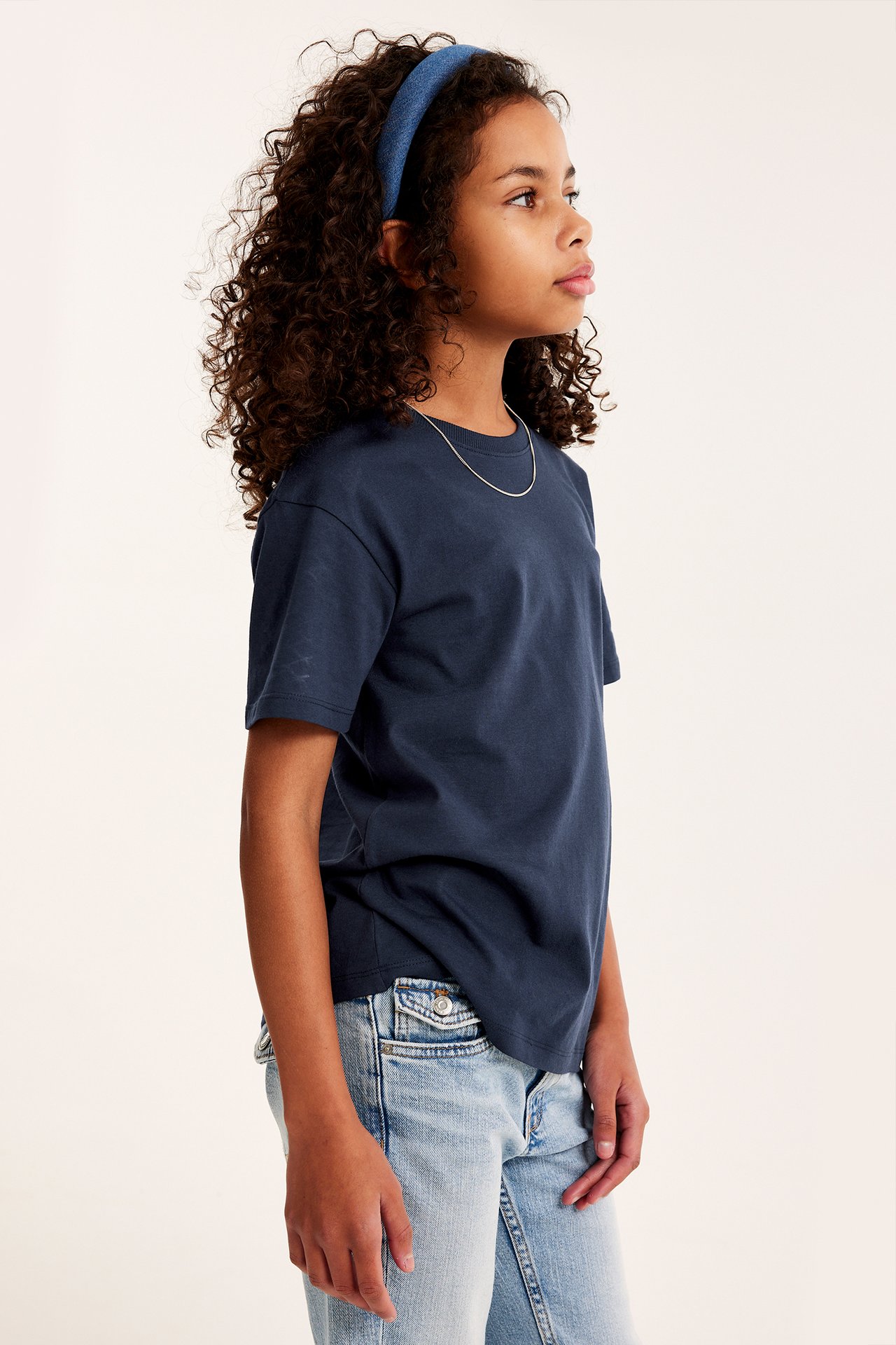 Sidovy: Ett barn i mörkblå bomulls-t-shirt, ljusblå jeans, blått hårband och silverhalsband.