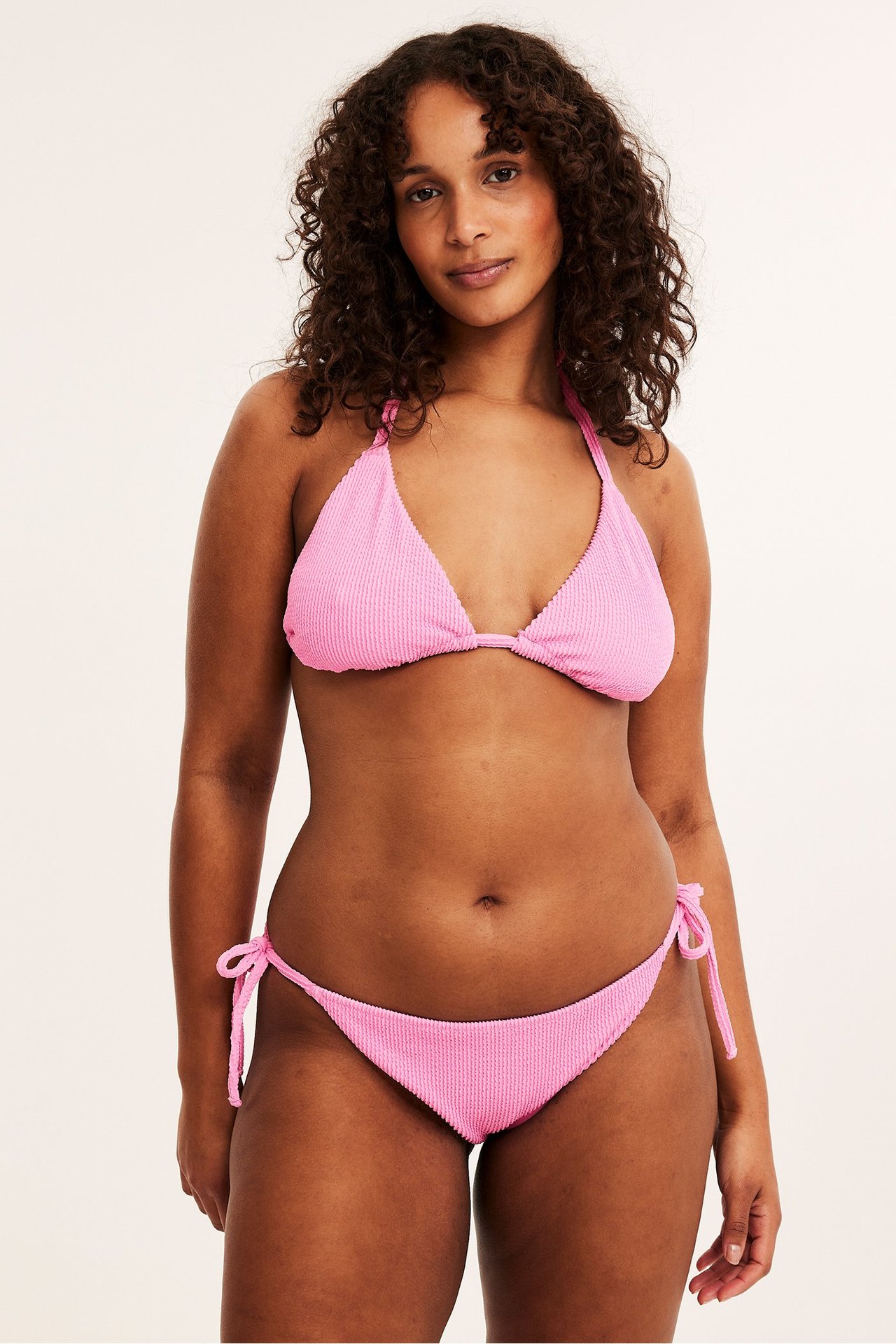 Widok z przodu: kobieta w jasnoróżowych, teksturowanych figach bikini brazilian z wiązaniami i pasującym topie.