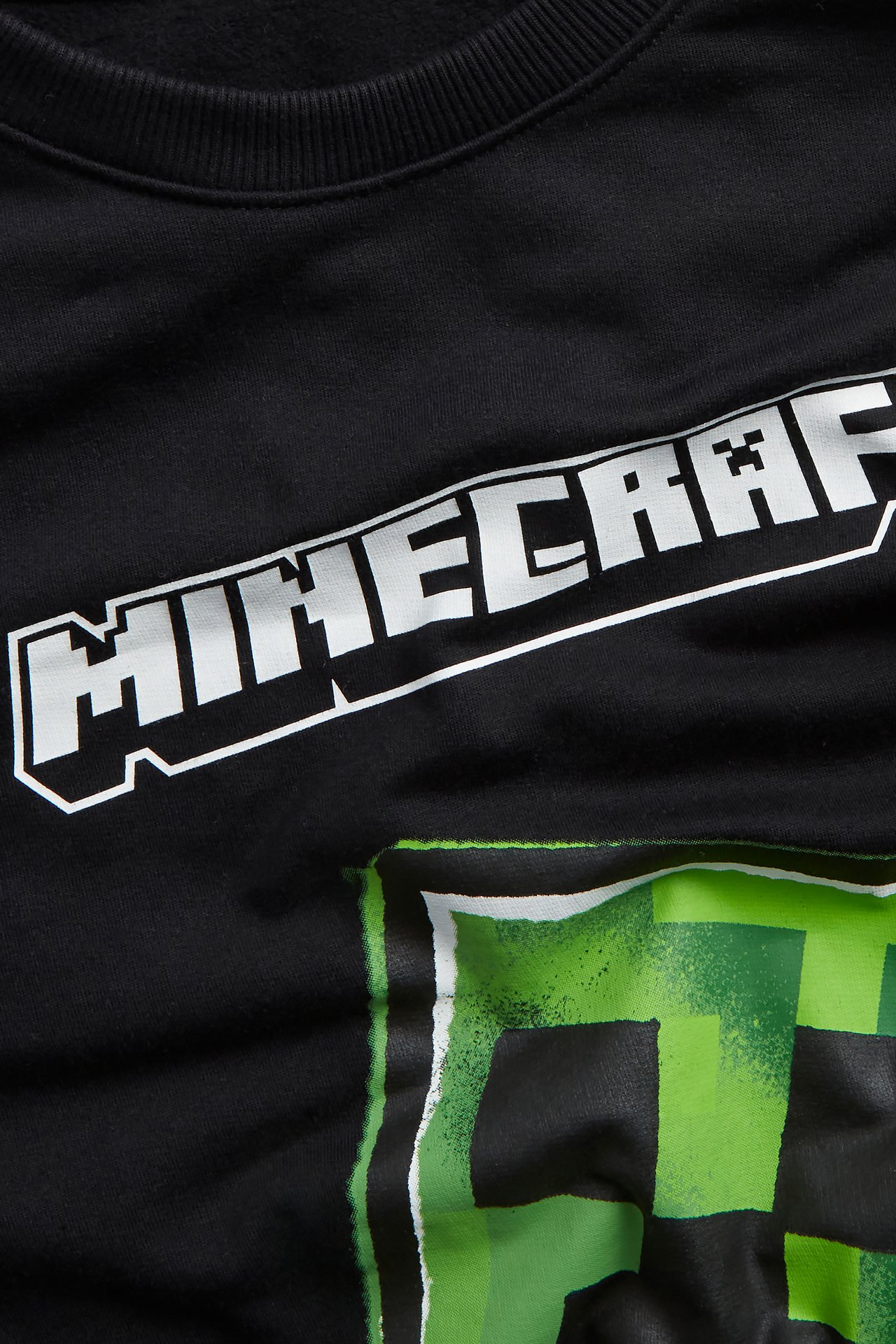 Lähikuva mustasta puuvillapaidasta, jossa valkoinen Minecraft-logo ja vihreä Creeper-painatus.