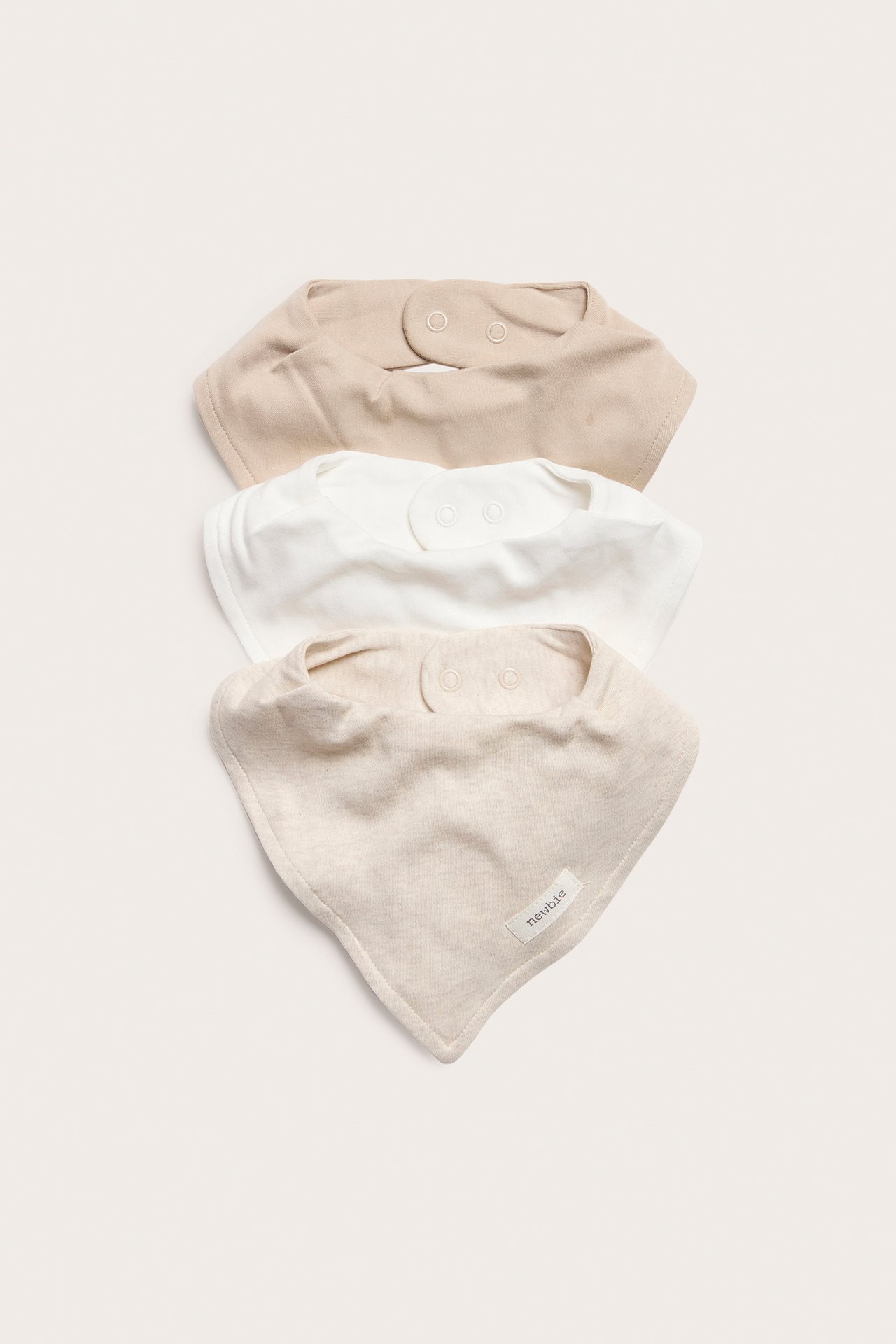 Uppifrån vy av tre enfärgade babyhaklappar: ljusbeige, vit och beige melange, med tryckknappar.
