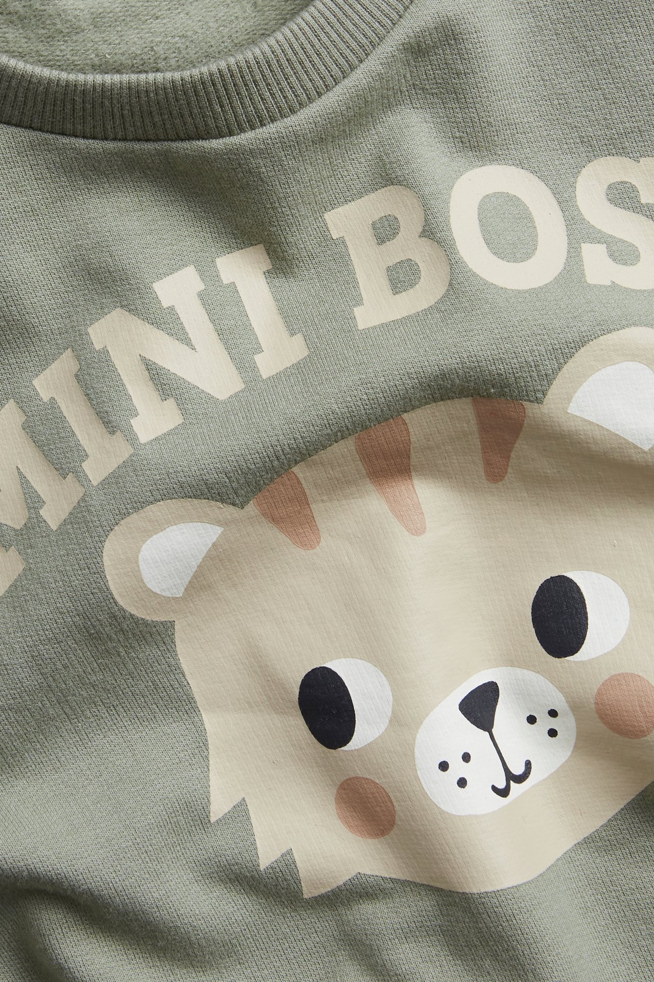 Lähikuva vauvan tummanvihreästä puuvillapaidasta, jossa on 'MINI BOSS' -teksti ja sarjakuvaleijonan kuva.