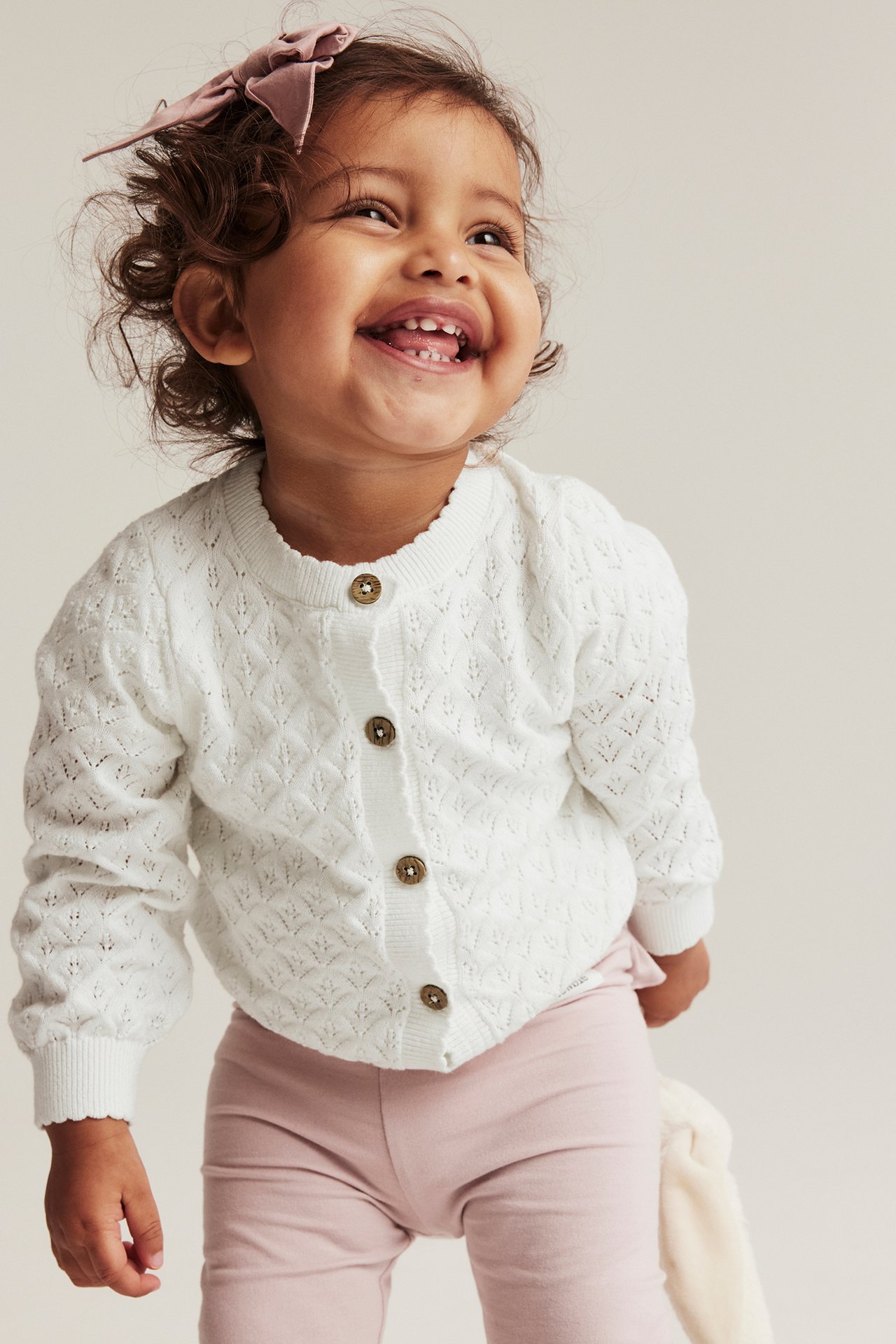 Framifrån: Leende baby i benvit mönsterstickad bomullskofta med knappar, ljusrosa leggings och rosa rosett.