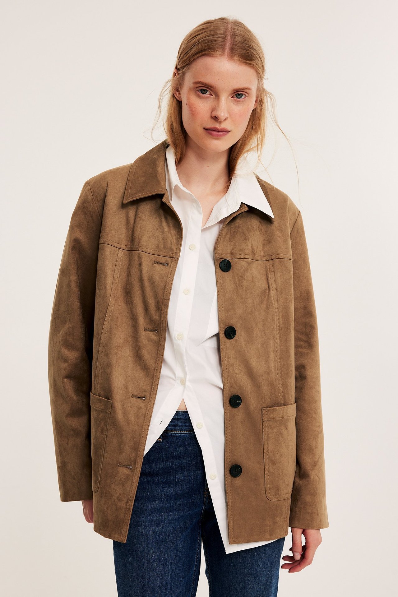 Framifrån, kvinna i brun fuskmockajacka med svarta knappar över vit skjorta och blå jeans.
