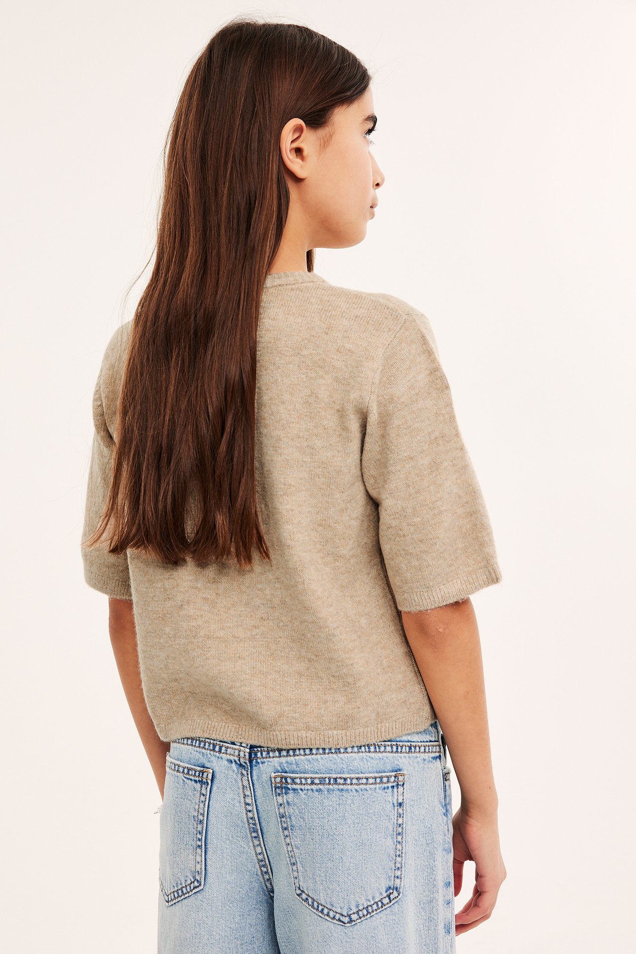 Bakfra av et barn i en beigemelert kortermet strikket cardigan og lyseblå jeans.