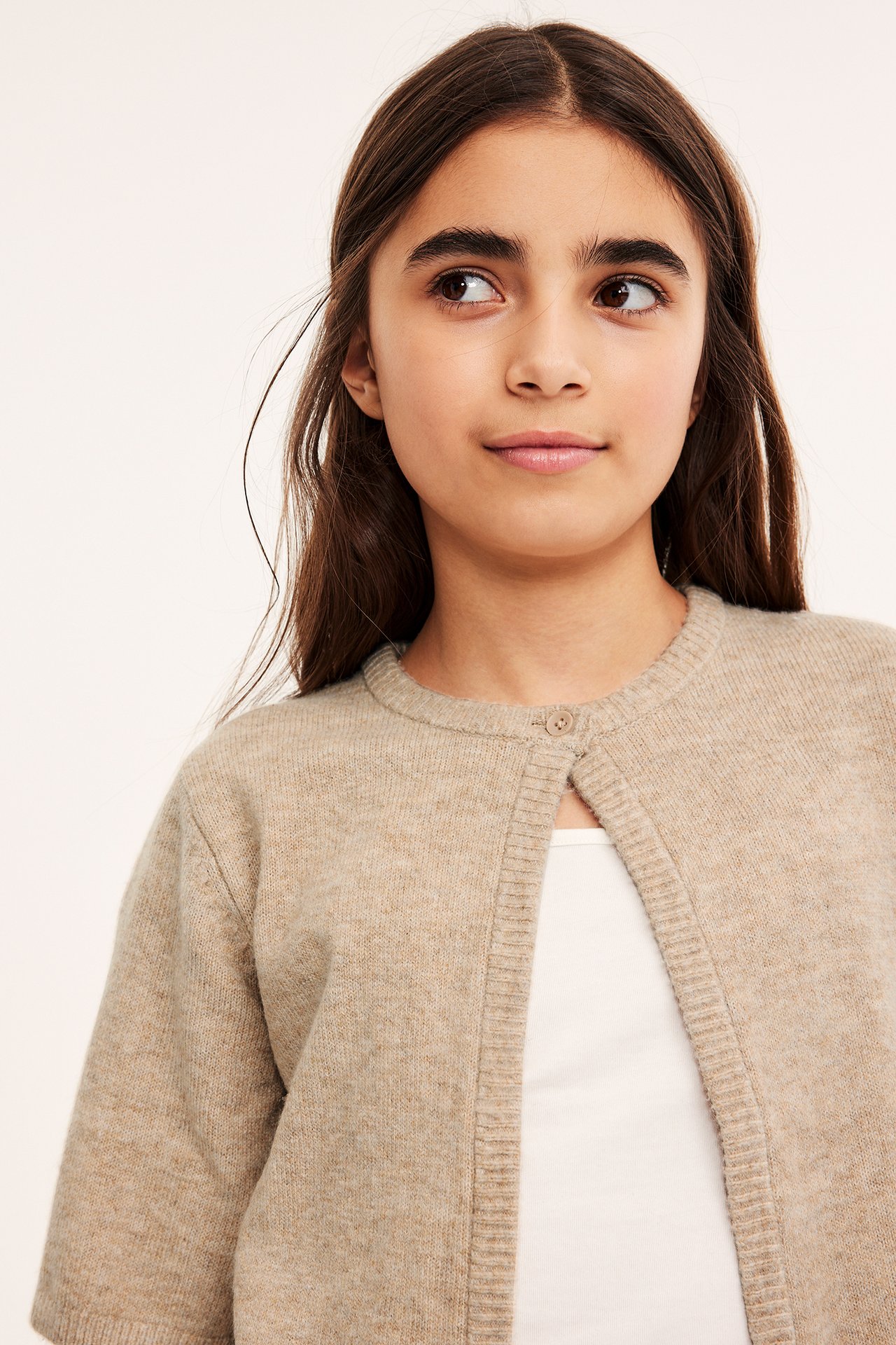 Frontbilde av et barn i en beigemelert kortermet strikket cardigan med én knapp, over en hvit topp.