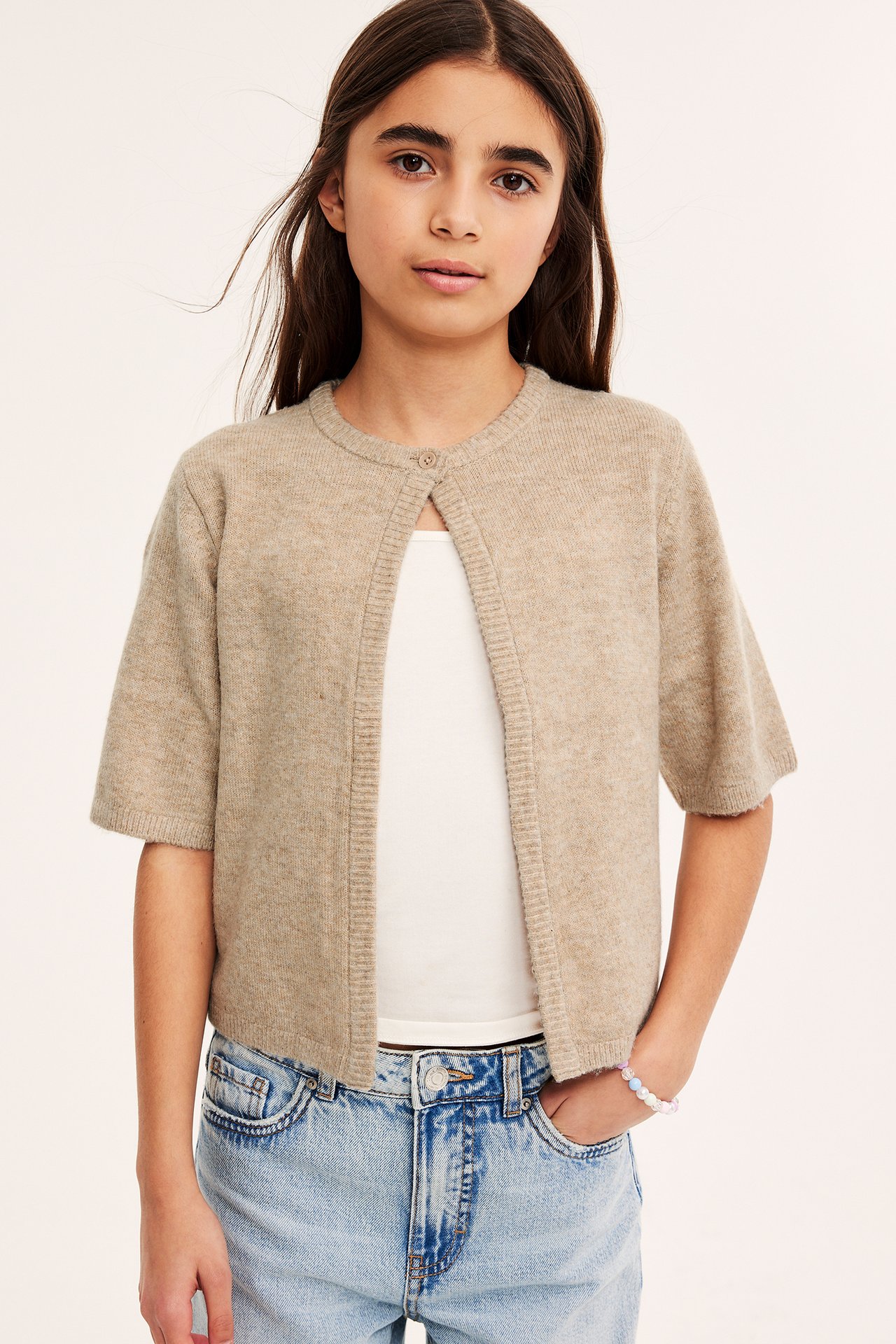 Barn iført en beige kortermet strikket cardigan med knapp, over hvit topp og lyseblå jeans.