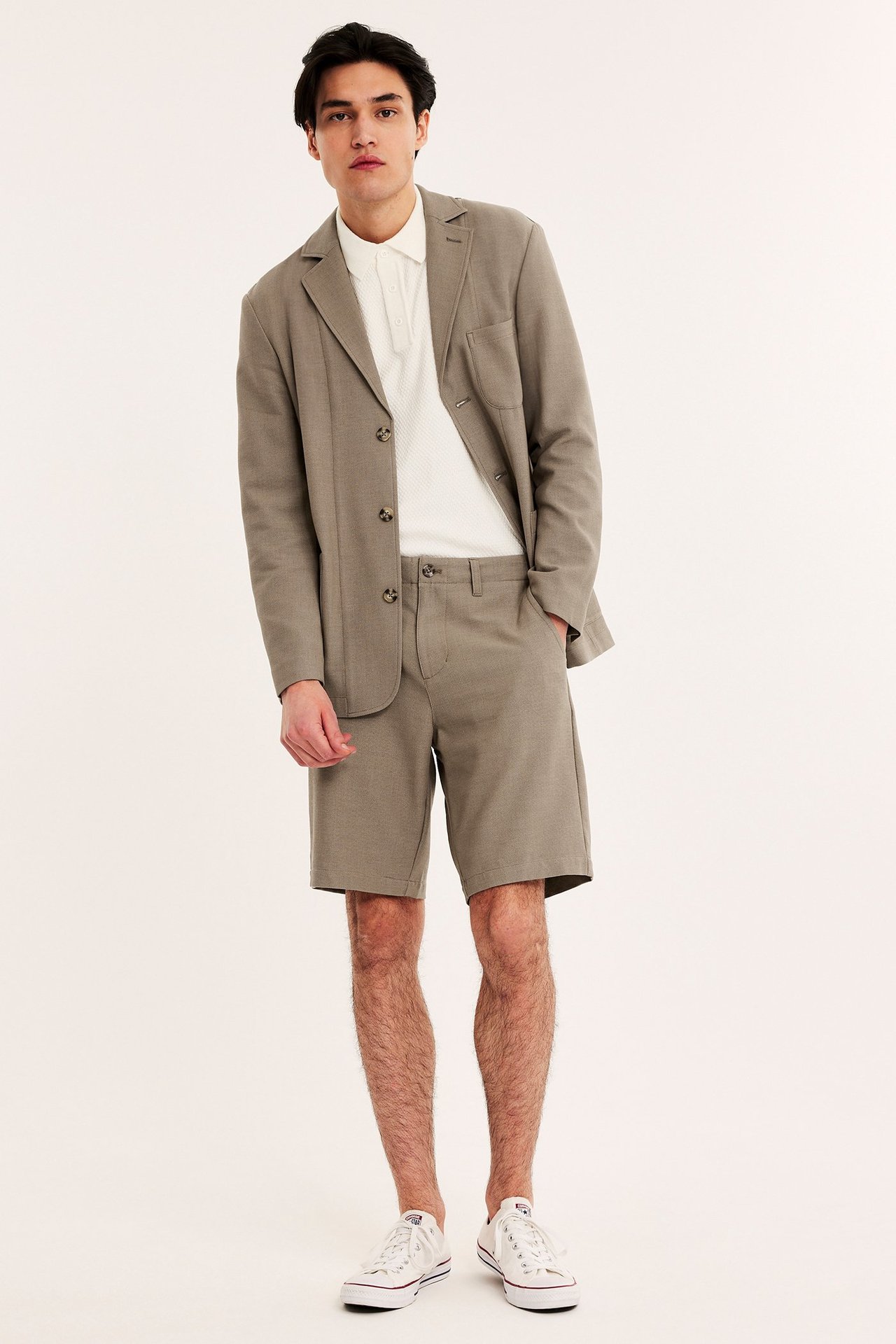 Edestäpäin mies yllään ruskeat joustavat chinoshortsit, yhteensopiva bleiseri, valkoinen pikeepaita ja valkoiset tennarit.
