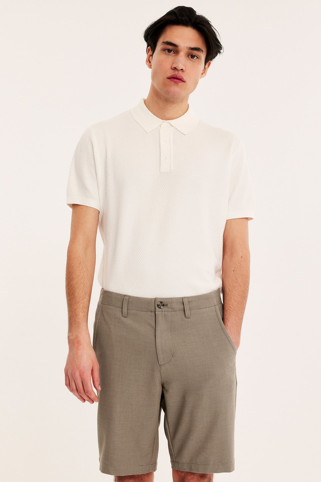 Edestä: mies ruskeissa chinoshortseissa taskuilla ja valkoisessa kuvioidussa lyhythihaisessa pikeepaidassa.