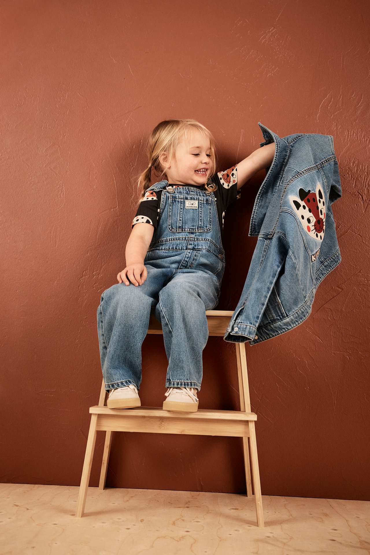 Framifrån: Barn bär ljusblå denimhängselbyxor, en svart djurmönstrad t-shirt, håller en denimjacka.