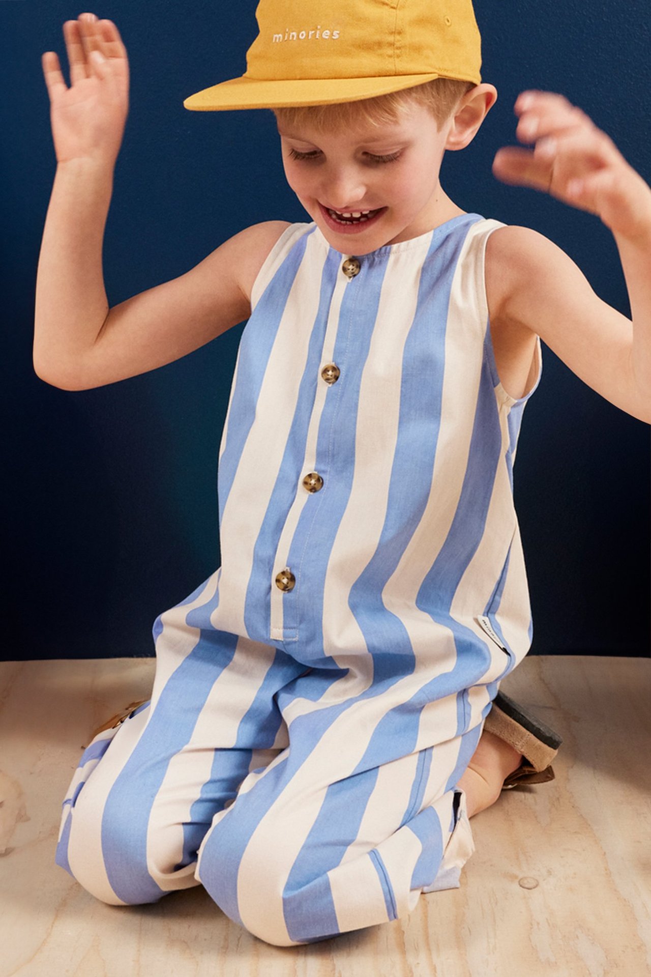 Widok z przodu: klęczące dziecko w niebiesko-białym bawełnianym kombinezonie w paski z guzikami i żółtą czapką.
