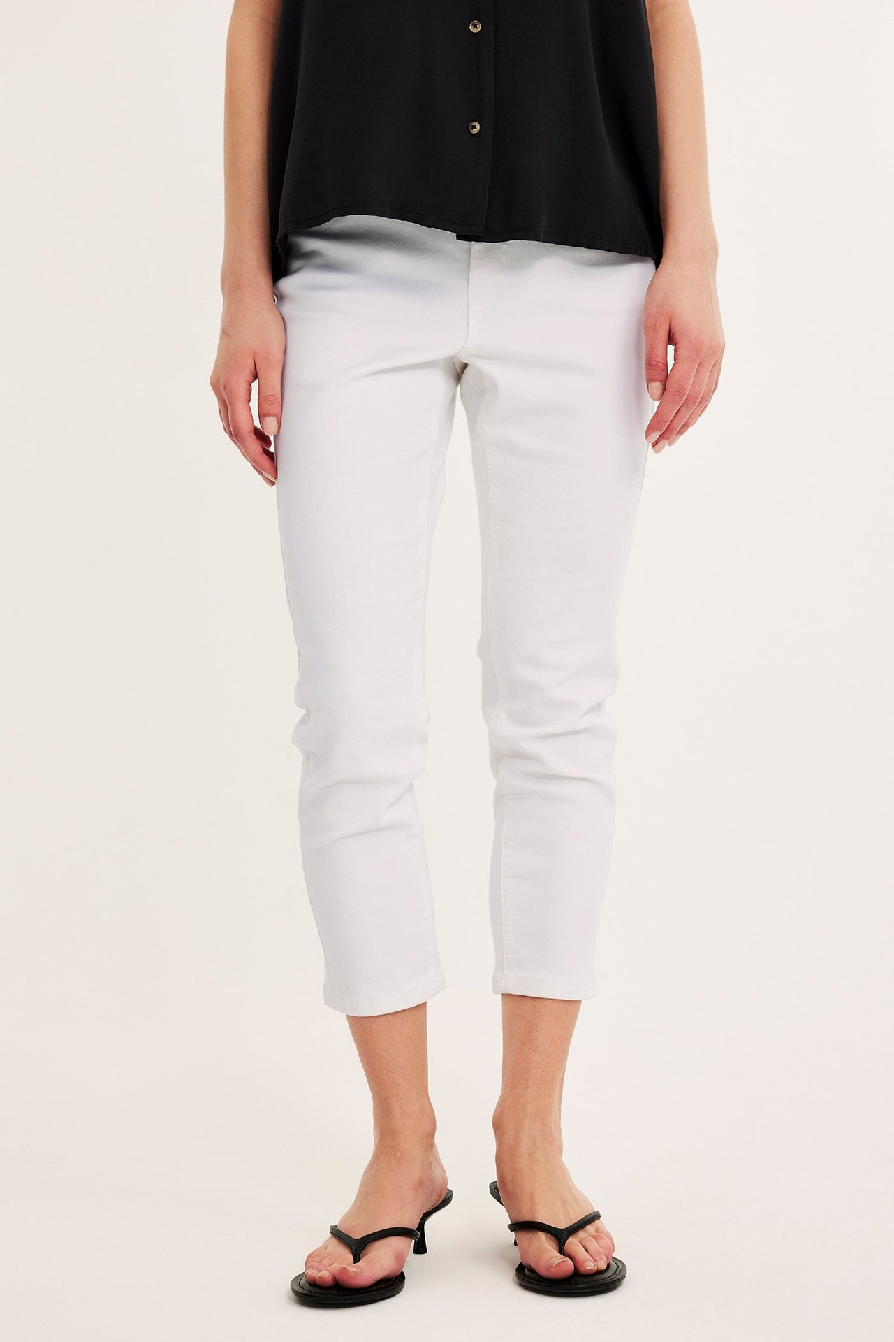 Framifrån, kvinna i vita croppade jeans med hög midja, svart ärmlös topp och svarta flip-flops med klack.