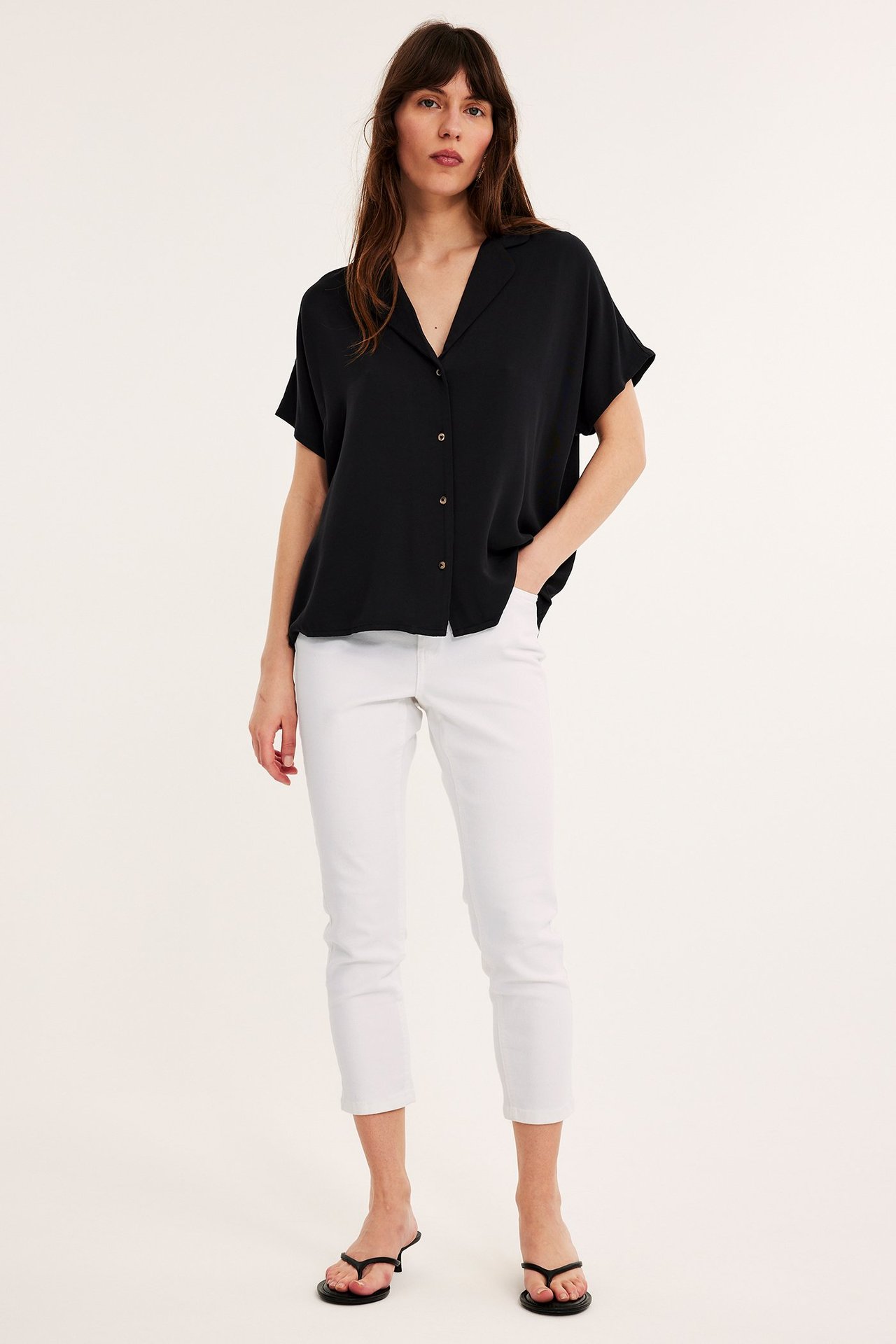 Framifrån: kvinna i vita croppade slim-fit jeans med hög midja, svart kortärmad skjorta och svarta flip-flops.