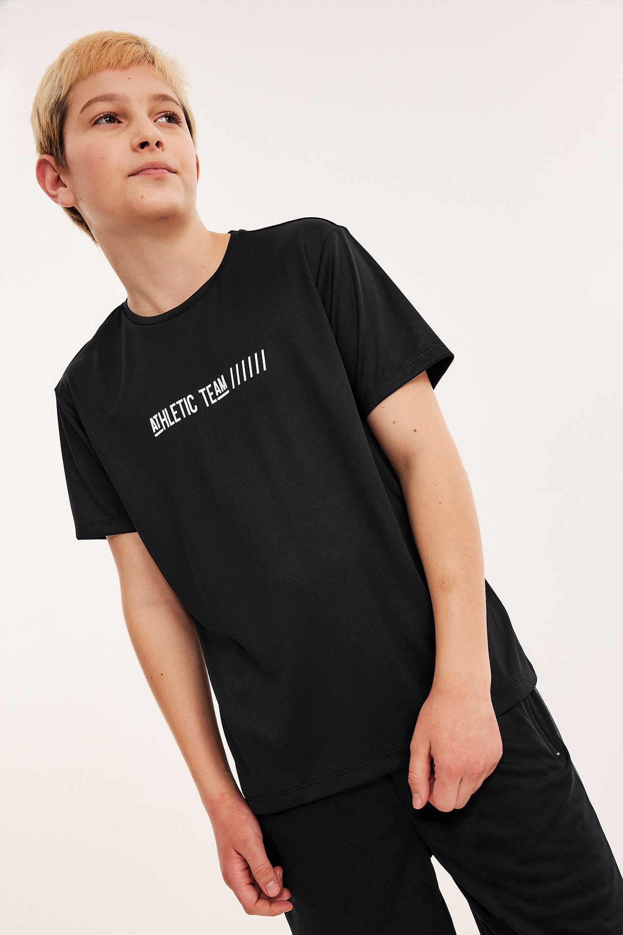 Widok z przodu: Dziecko w czarnym poliestrowym T-shircie z białym napisem 'ATHLETIC TEAM', czarne szorty.