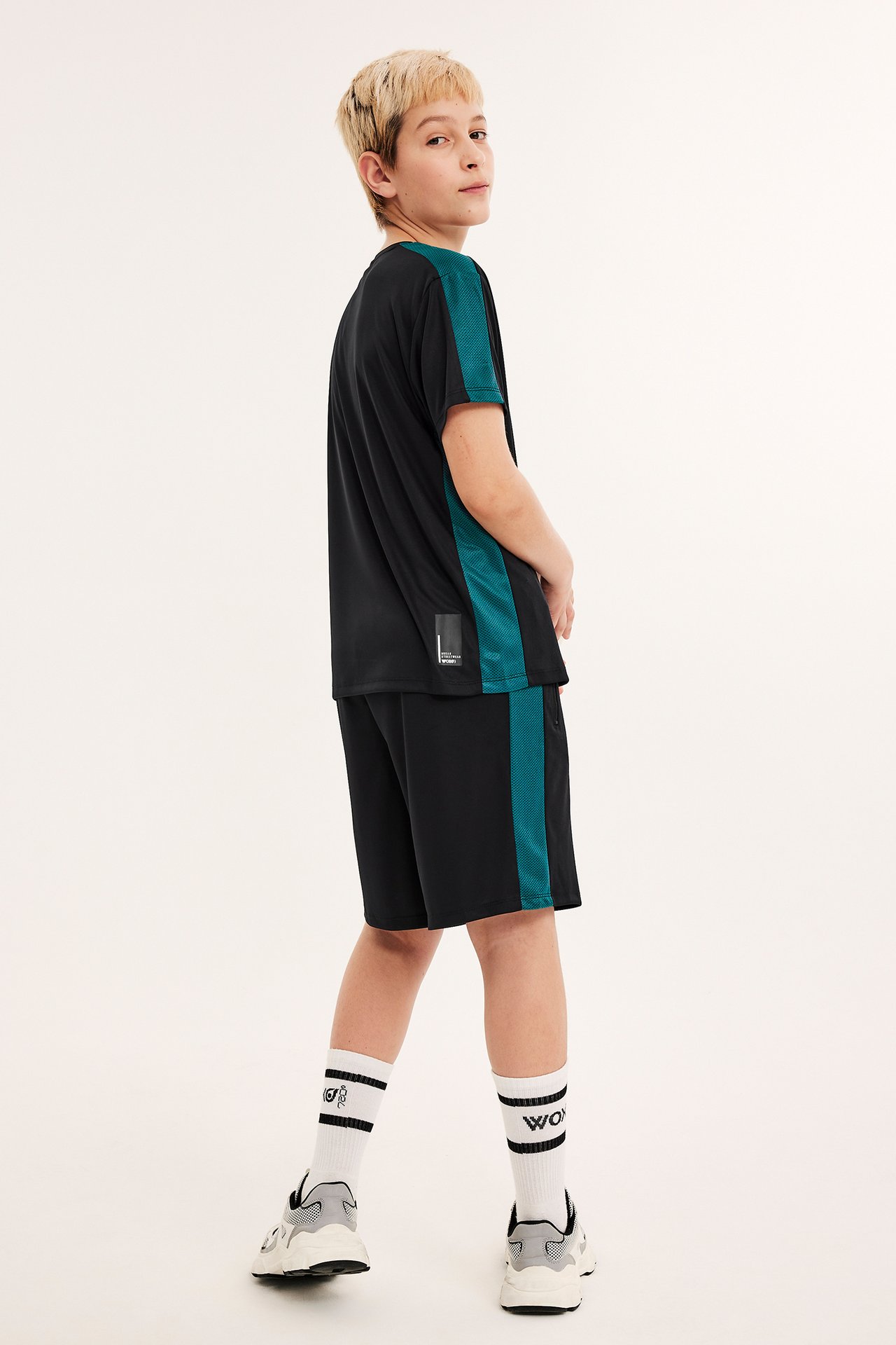 Bakifrån: ett barn i svarta träningsshorts med turkos mesh-rand, matchande t-shirt, vita strumpor och sneakers.