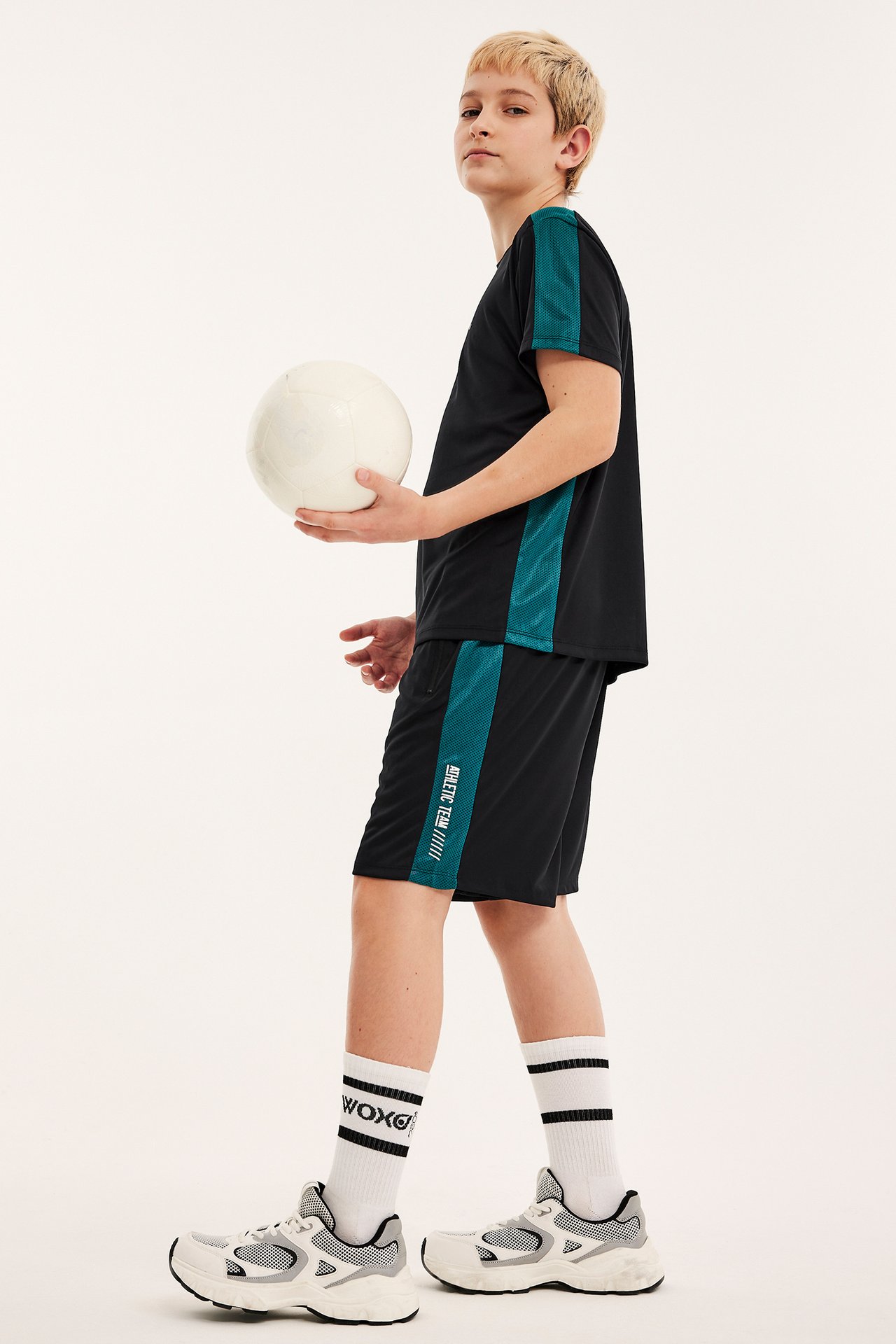 Sidovy: Barn i svarta shorts med turkos mesh, matchande t-shirt, vita strumpor, sneakers, håller en boll.
