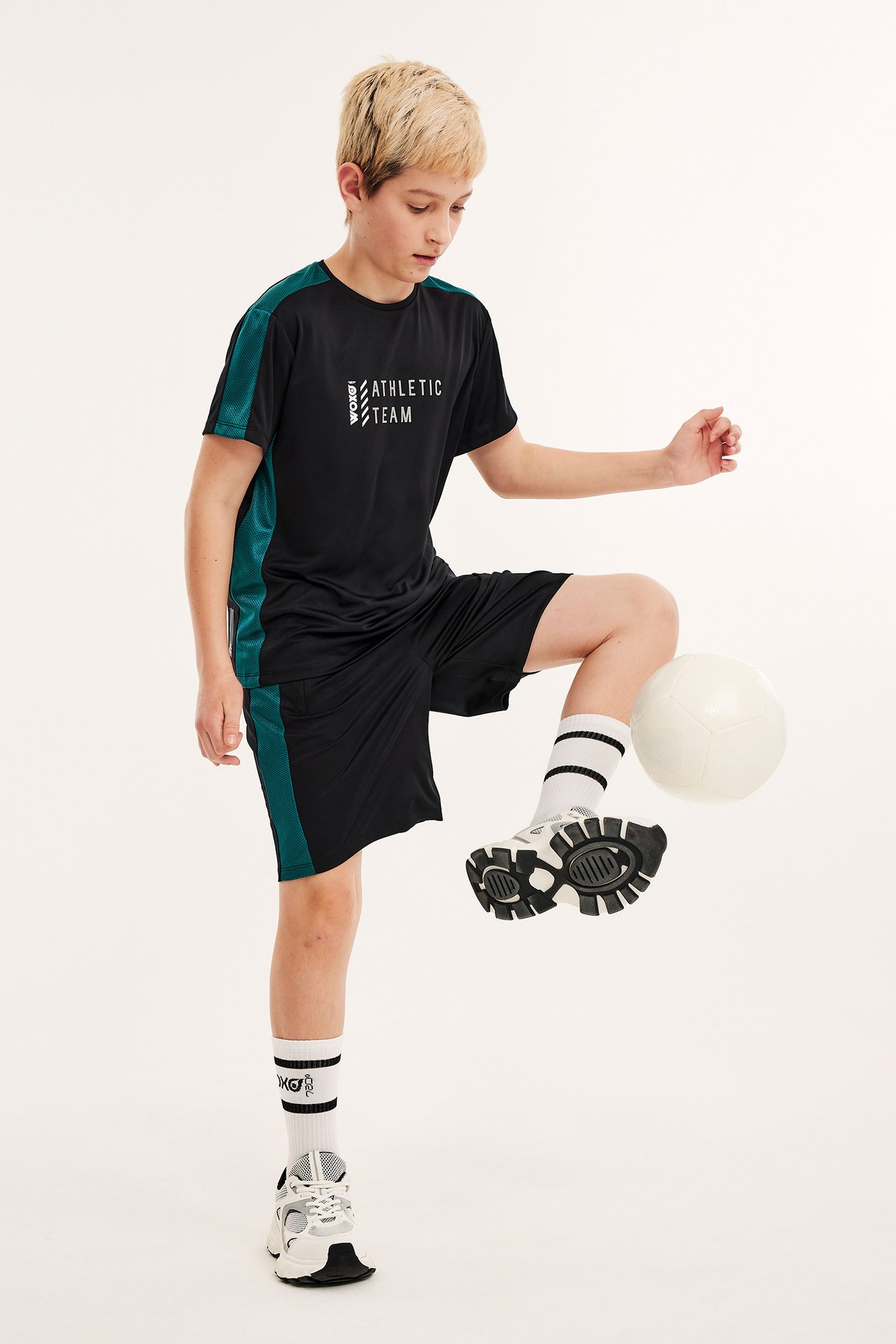 Framifrån: Barn i svarta träningsshorts med grönt nät, svart t-shirt, vita strumpor och sneakers.