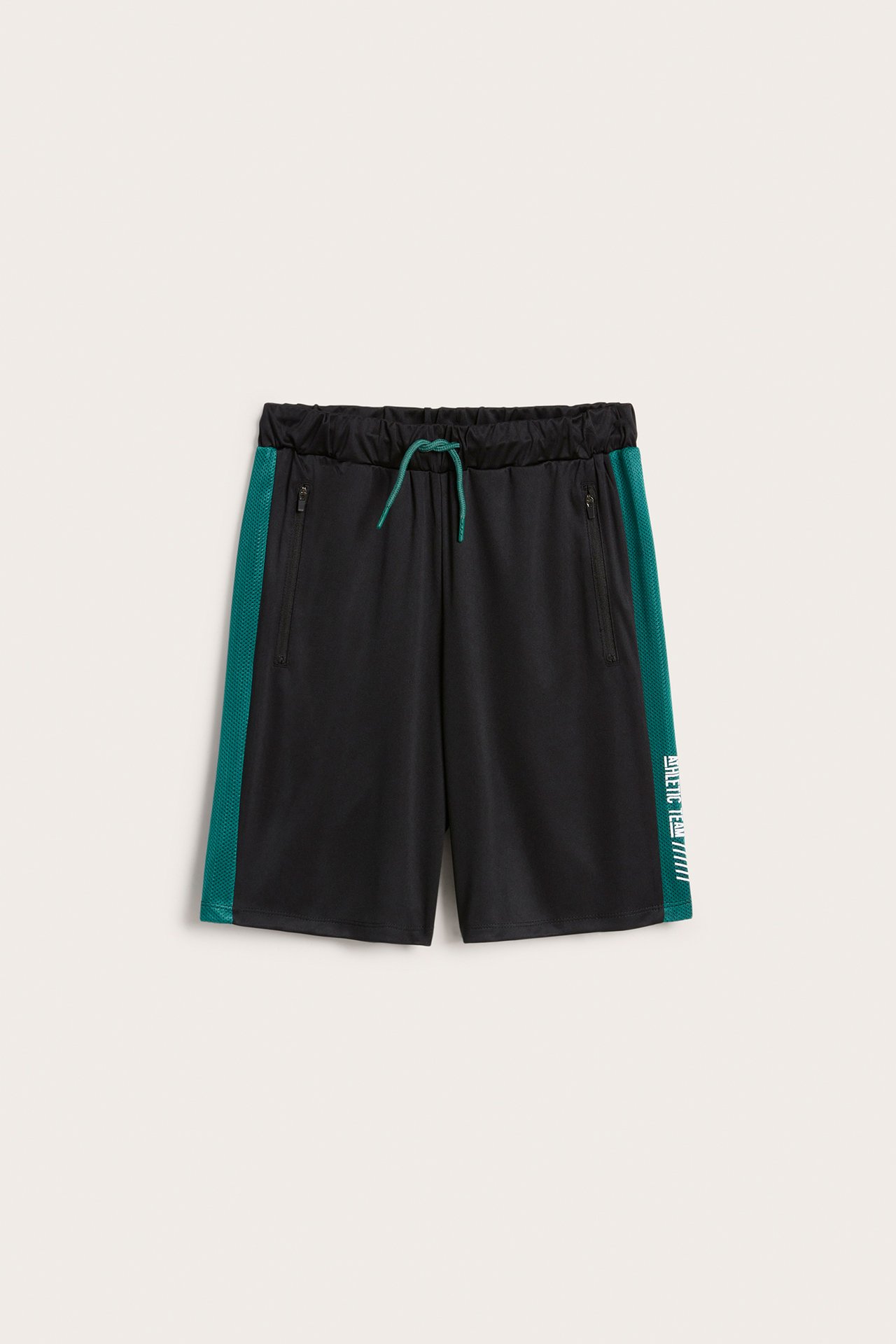 Frontvy: Svarta polyesterträningsshorts med gröna meshränder, vit text, grön dragsko och dragkedjefickor.