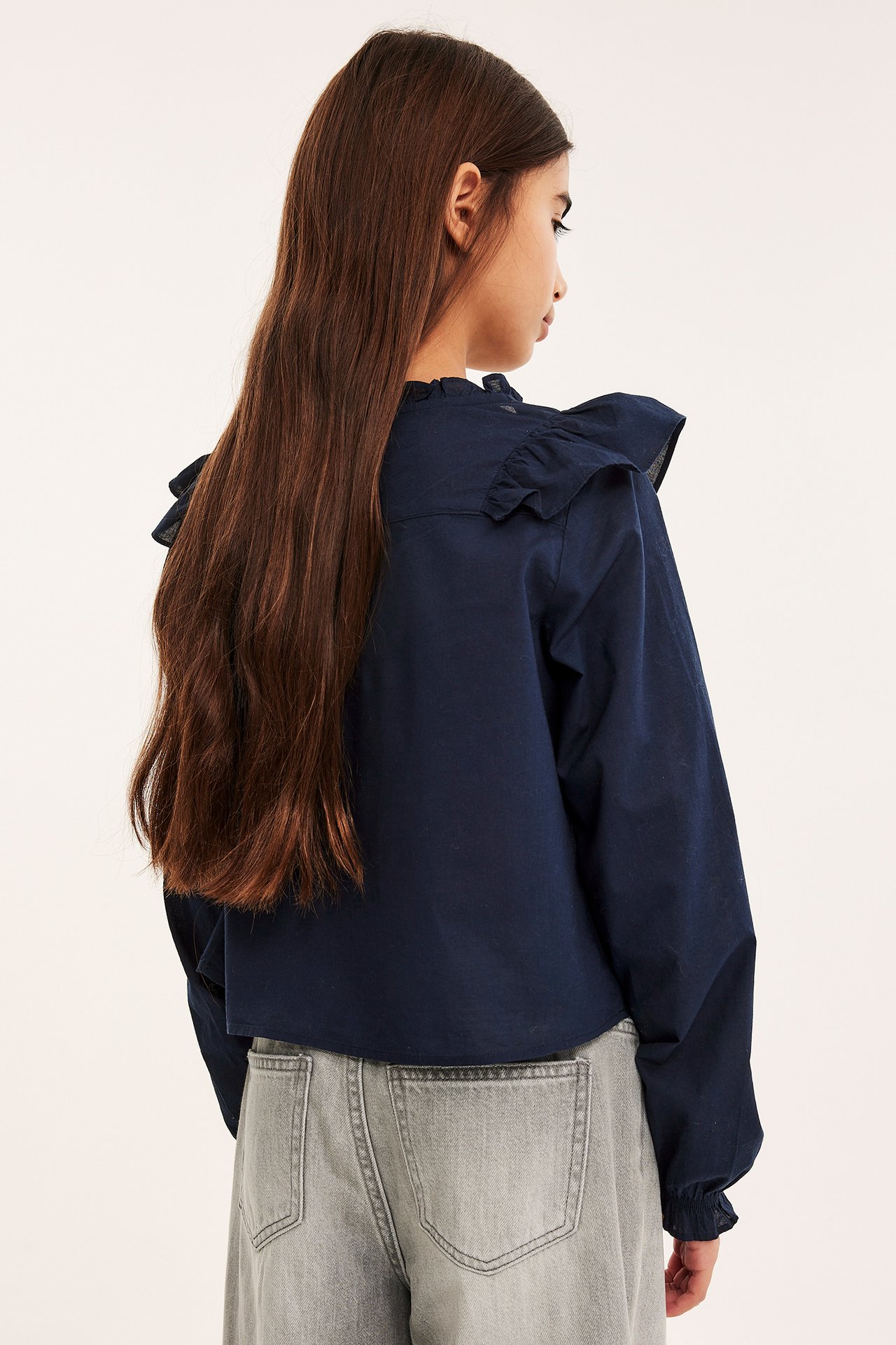 Bakfra, et barn i marineblå bomullsbluse med volanger og lysegrå vide jeans.