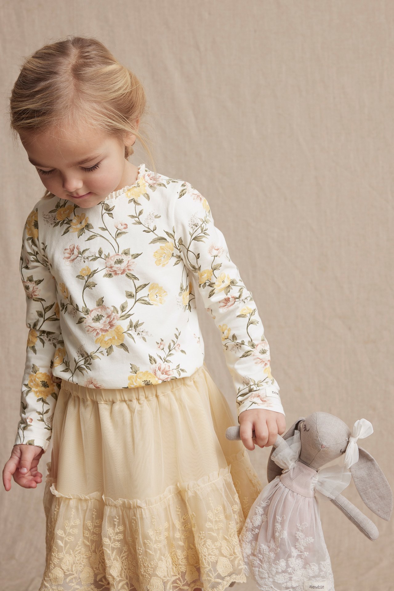 Forfra: Barn i offwhite blomstret jerseytop med blondekant, beige skjørt, holder kanindukke.