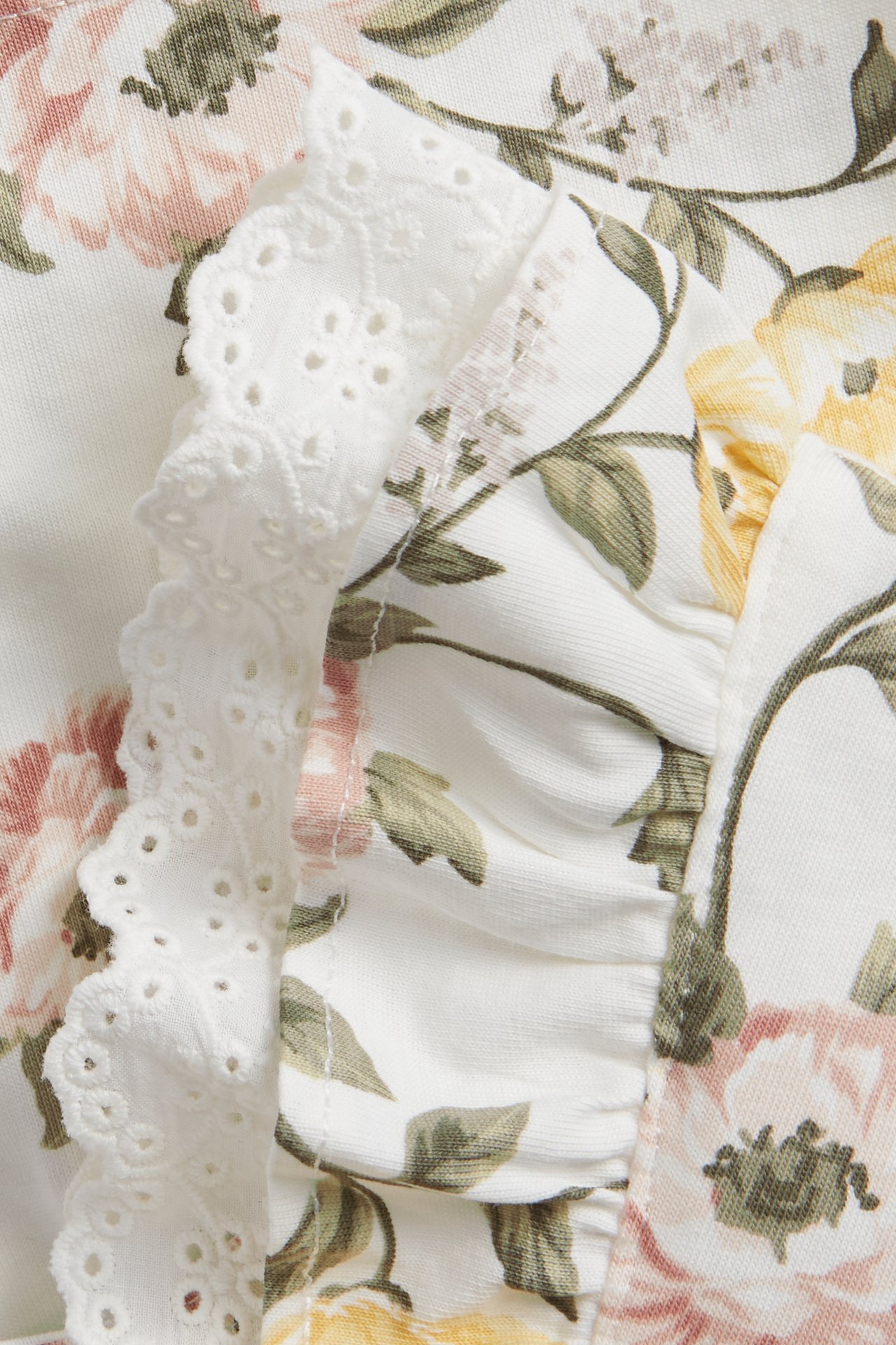 Nærbilde av en offwhite blomstret bomullskjole med rosa og gule blomster, med hvit blonde- og stoffvolang.