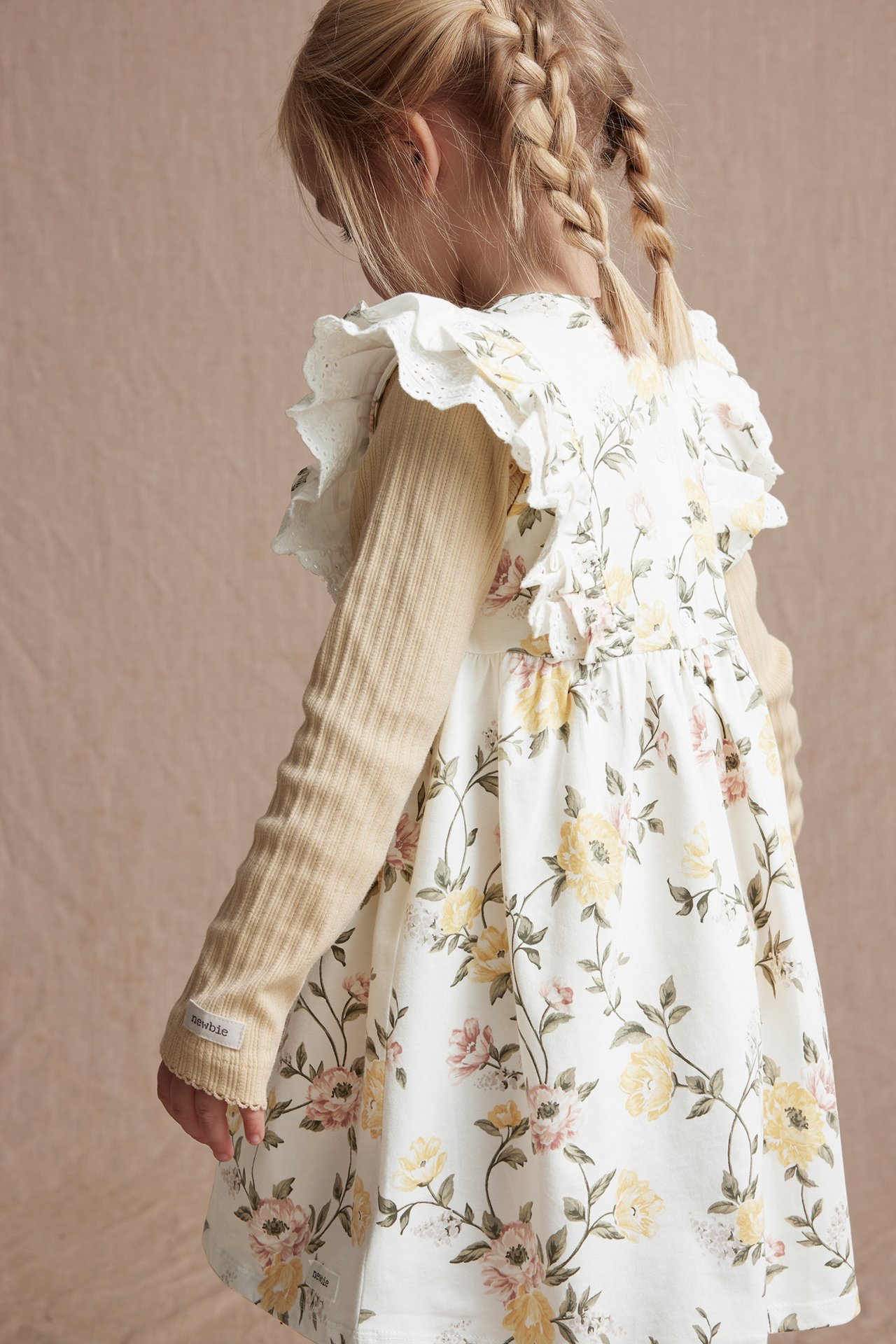 Barn, snett bakifrån: Offwhite blommig klänning med volanger över en beige ribbad långärmad topp.