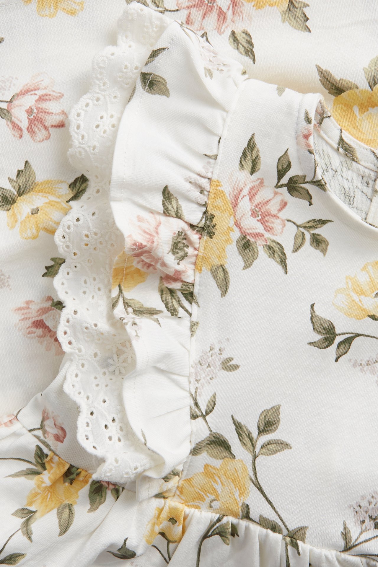 Närbild av en offwhite bomullsjerseyklänning med gult och rosa blommönster och volangspetsdetaljer.