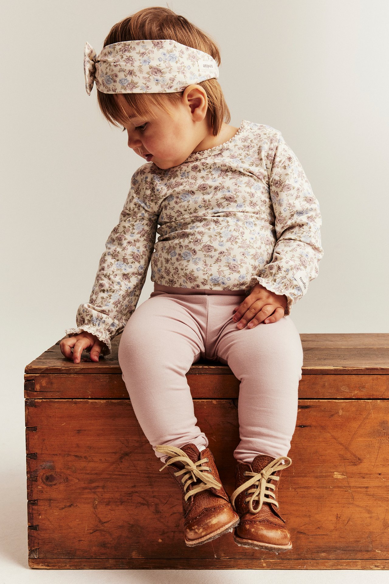 Framifrån: baby i ljusrosa leggings, blommig topp, matchande pannband och bruna snörskor.