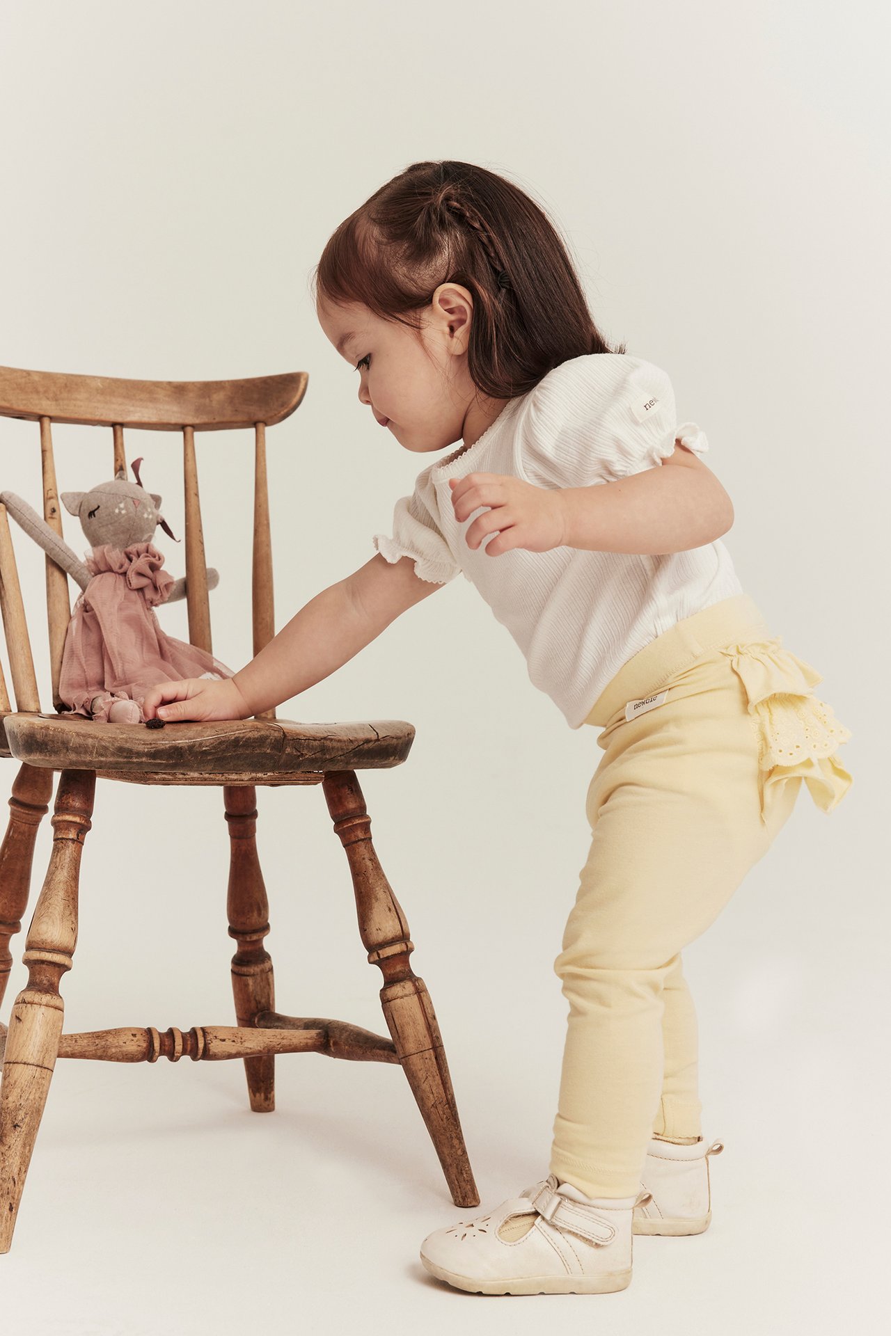 Sidovy av en babyflicka i gula leggings med volanger bak, en vit ribbad topp och ljusbeige skor.