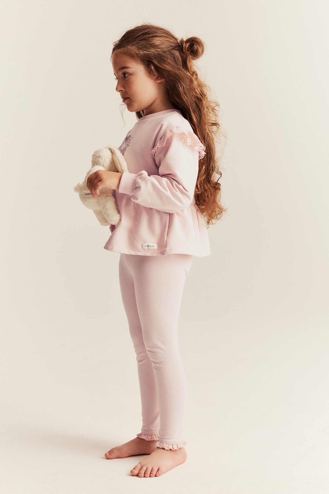 Widok z boku dziecka w jasnoróżowych legginsach z koronką i pasującym topie, trzymającego białego pluszaka.