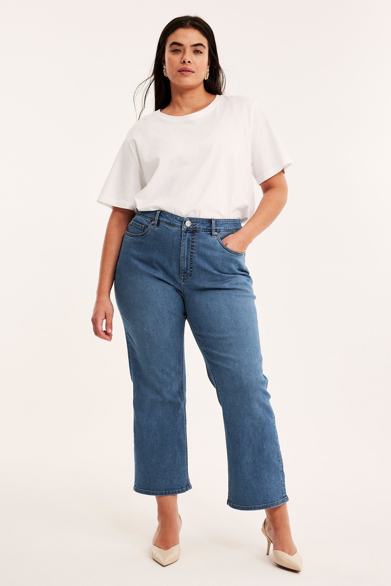 Frontvy av en kvinna i blå höga cropped flare jeans, en vit t-shirt, beiga klackar och guldörhängen.