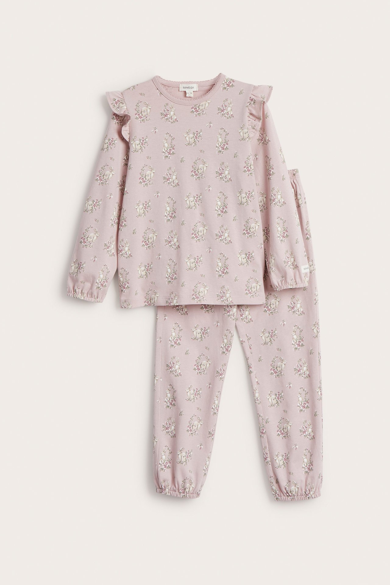 Edestä: Lasten vaaleanpunainen puuvillapyjama, jossa olkapäässä röyhelöt ja kukka-kanikuvio.