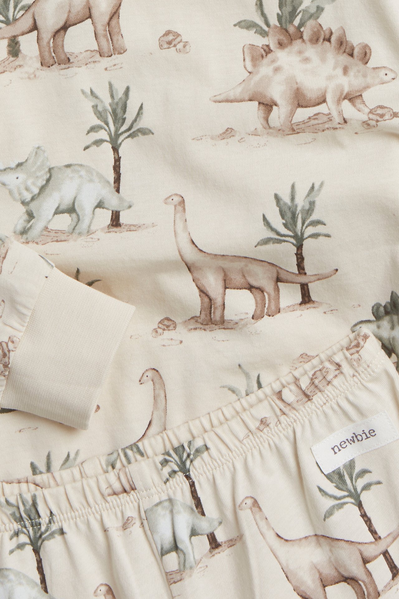 Närbild på en ljusbeige tvådelad pyjamas i trikå för barn, med dinosauriemönster och ribbade detaljer.