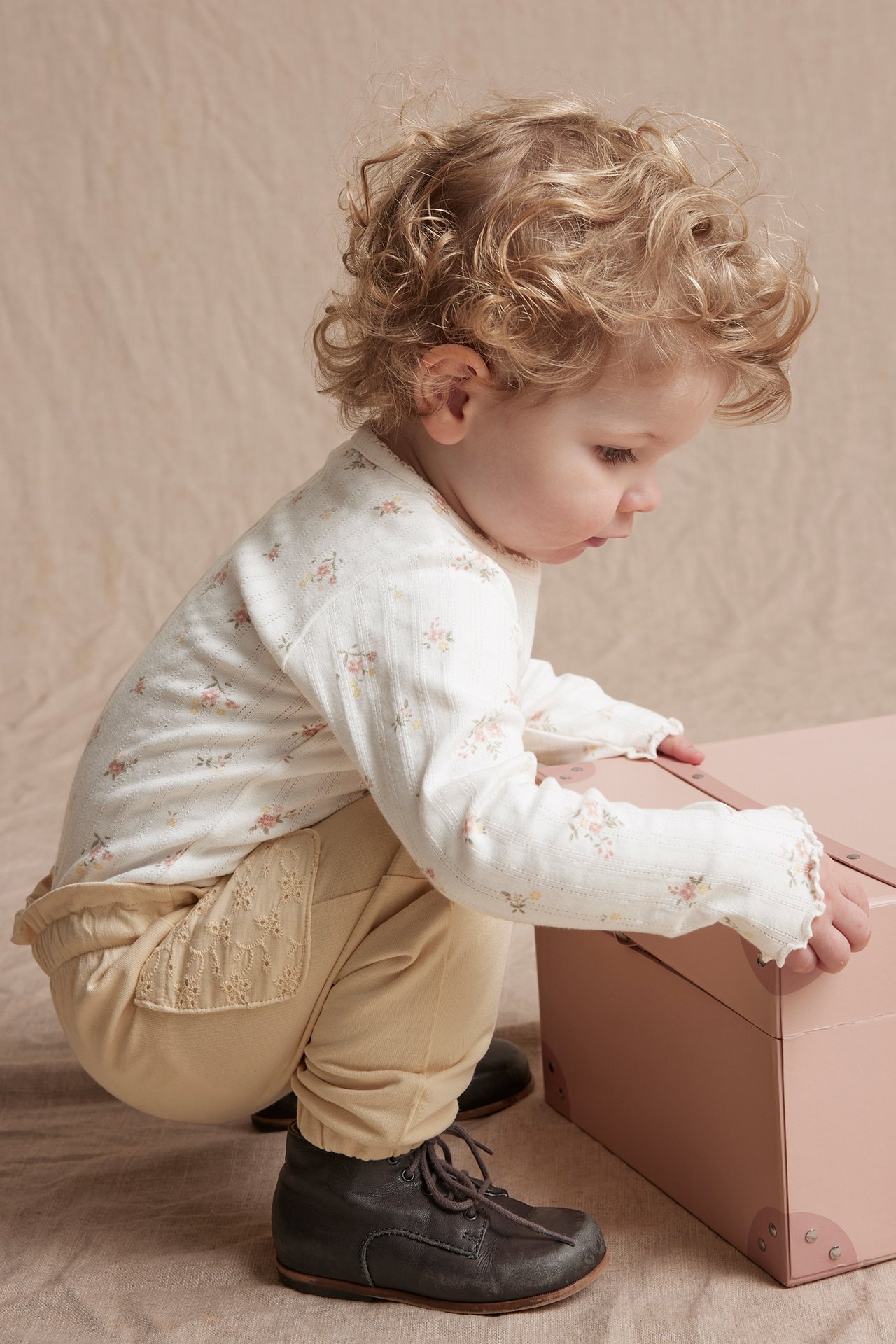 Sidebilde av baby i offwhite pointelle blomstret langermet body, beige bukser og mørkebrune sko.