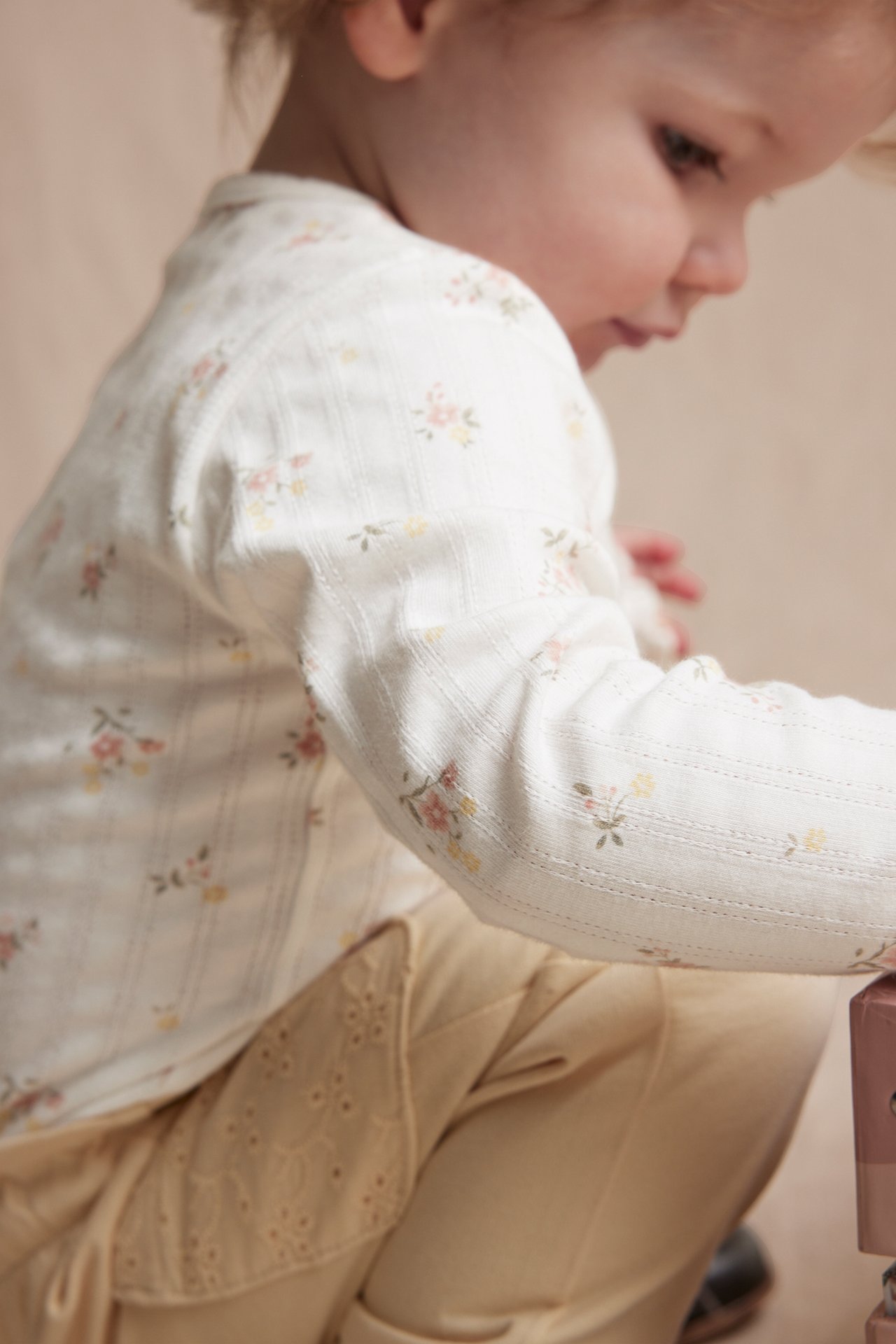 Sidebilde av baby i offwhite pointelle ribb langermet body med blomstertrykk og lysegule bukser.