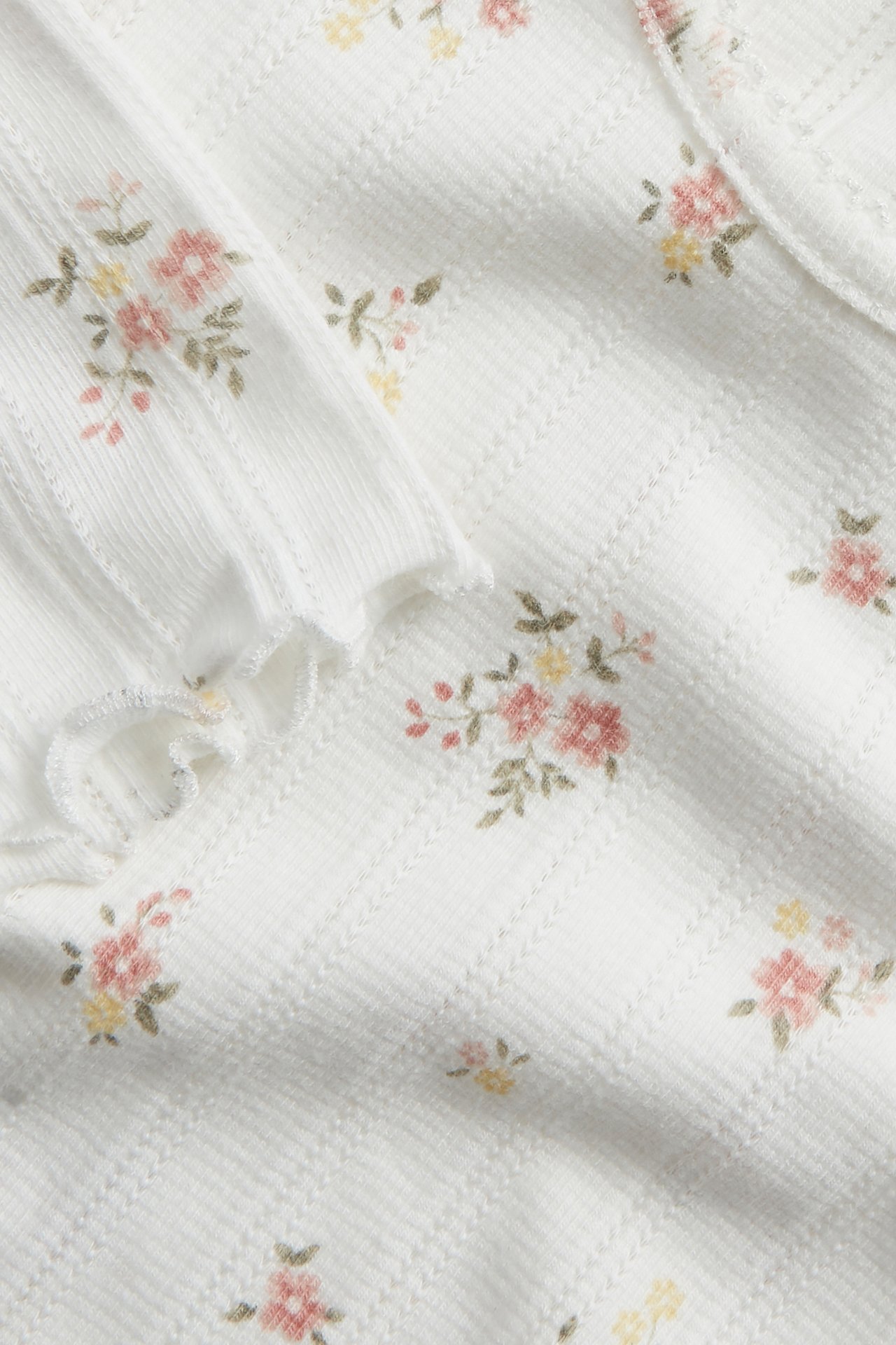 Nærbilde av en offwhite pointelle ribbet babybody med lang erm, blomstertrykk og bølgete kanter.