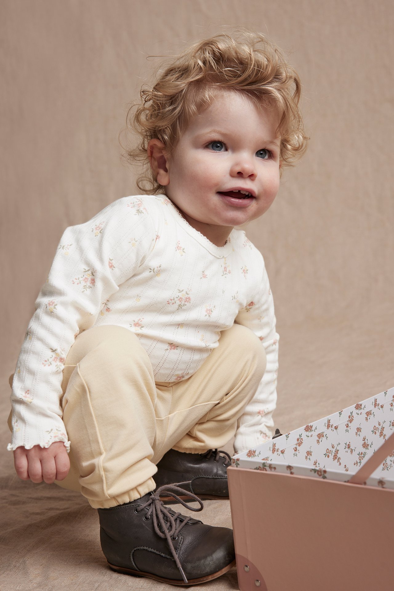 Frontvisning av baby i offwhite pointelle blomstret langermet body, lysegule bukser og mørkegrå sko.