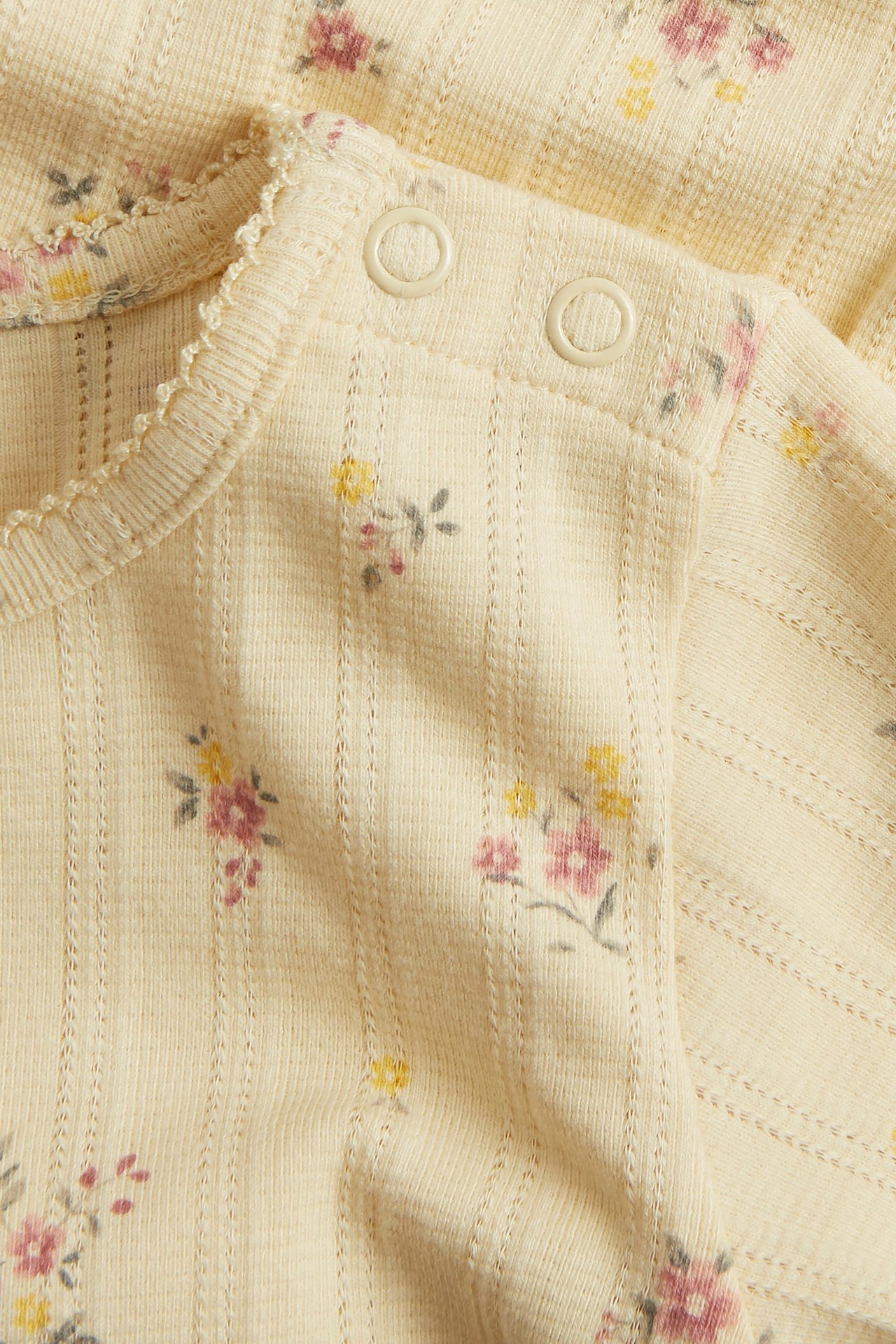 Nærbilde av en babygul pointelle ribbestrikket langermet body med blomstertrykk, bølgete kanter og trykknapper.