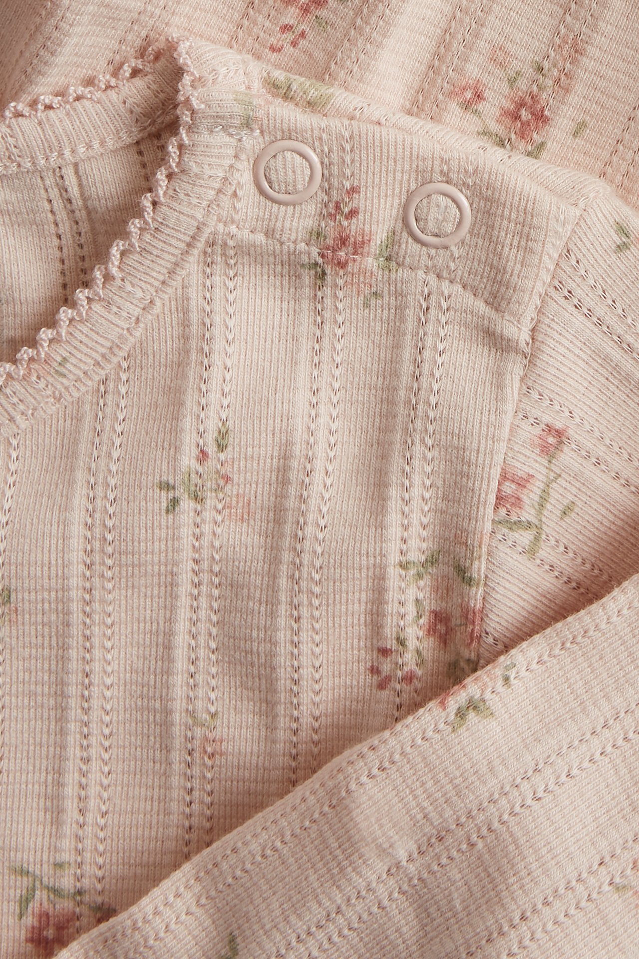 Nærbilde av en lyserosa pointelle ribbet babybody med blomstertrykk og trykknapper på skulderen.